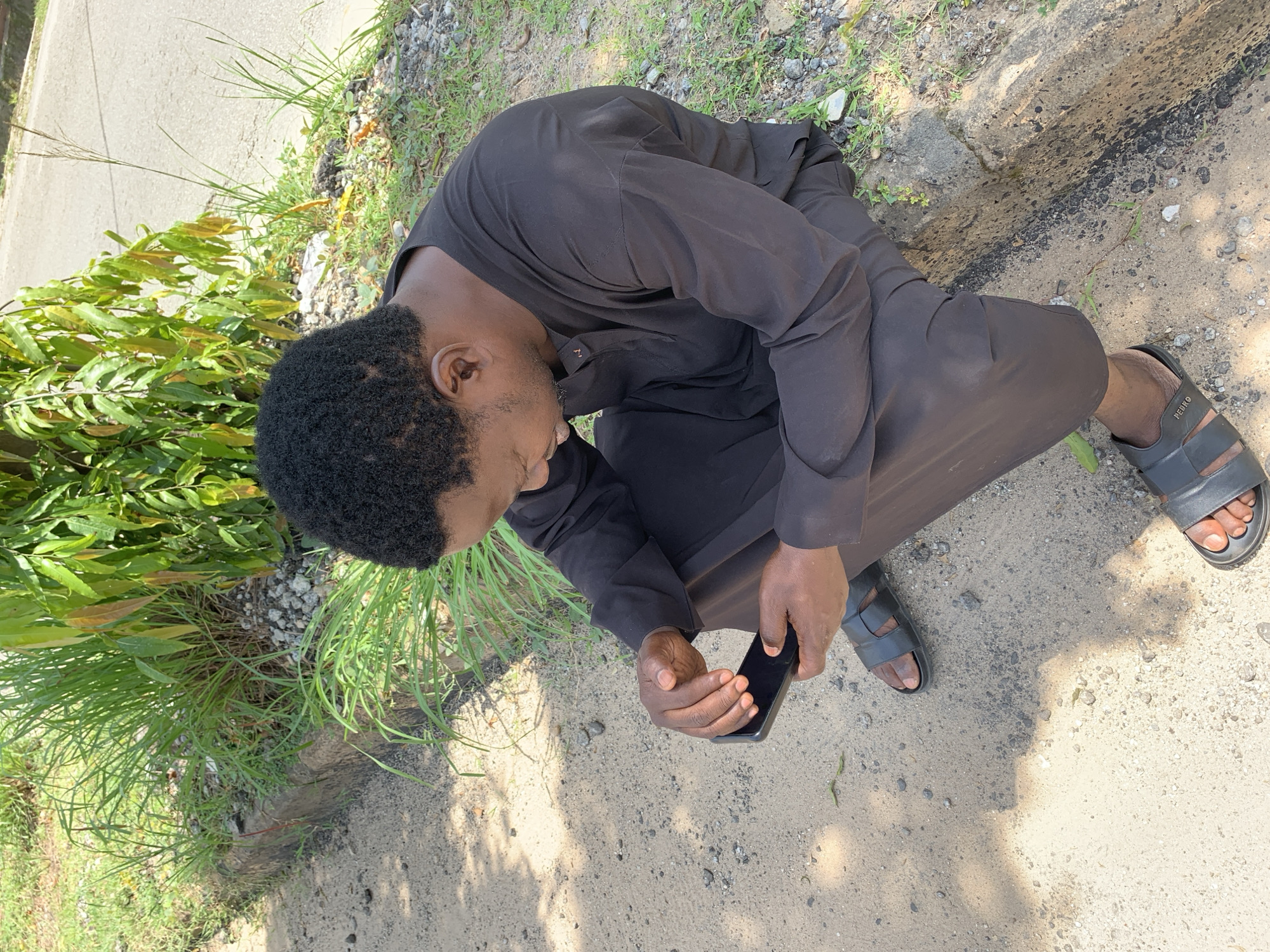 A person, appearing to be a young Black male, is crouched down outdoors, looking at a smartphone. He is wearing a dark, possibly black, tunic and dark sandals. The ground beneath him is a mix of dirt and small gravel, with patches of green grass and some leafy plants to his left. A road is visible in the background. The lighting suggests it is daytime, with shadows indicating sunlight. The scene appears casual and perhaps contemplative, given the subject's focus on the phone and his relaxed posture. No visible text is present on the clothing or objects. The image seems to capture a moment of everyday life in a rural or semi-rural setting, possibly in Galadimawa Village, Nigeria.