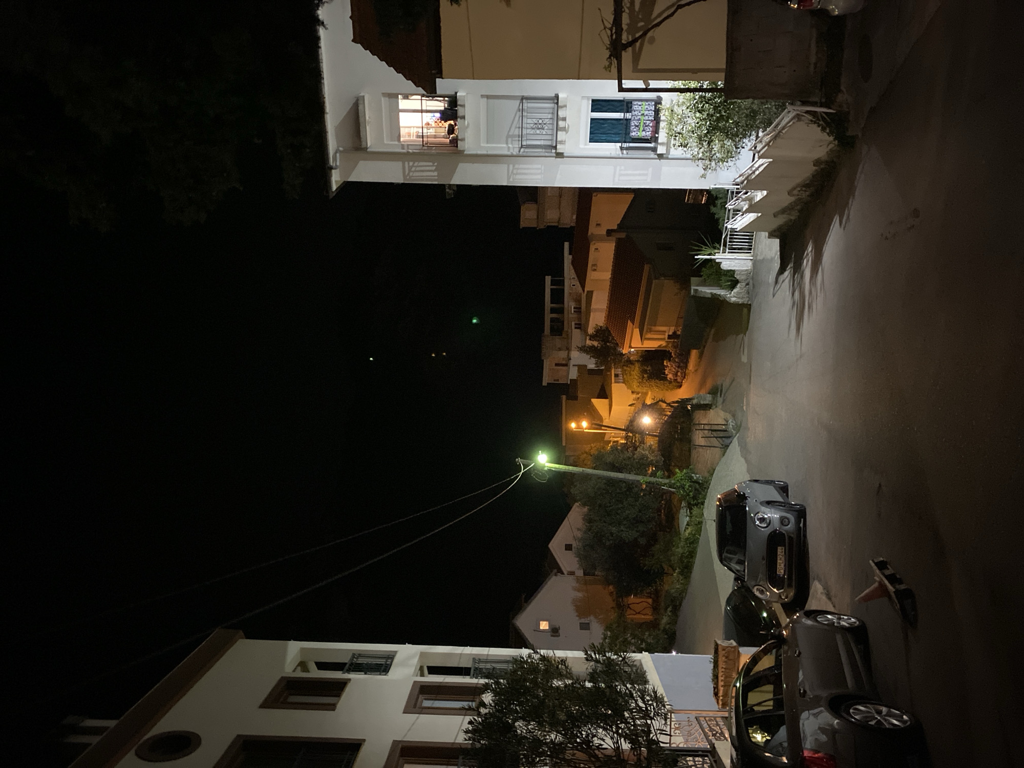 The image shows a street in Budva, Montenegro at night. The scene is illuminated by street lights and lights from buildings. There are several buildings of varying heights lining the street, some with balconies and windows lit up. Cars are parked along the street. There is a utility pole with wires running across the image. Vegetation such as trees and bushes are visible. An orange safety cone is on the street next to the cars.