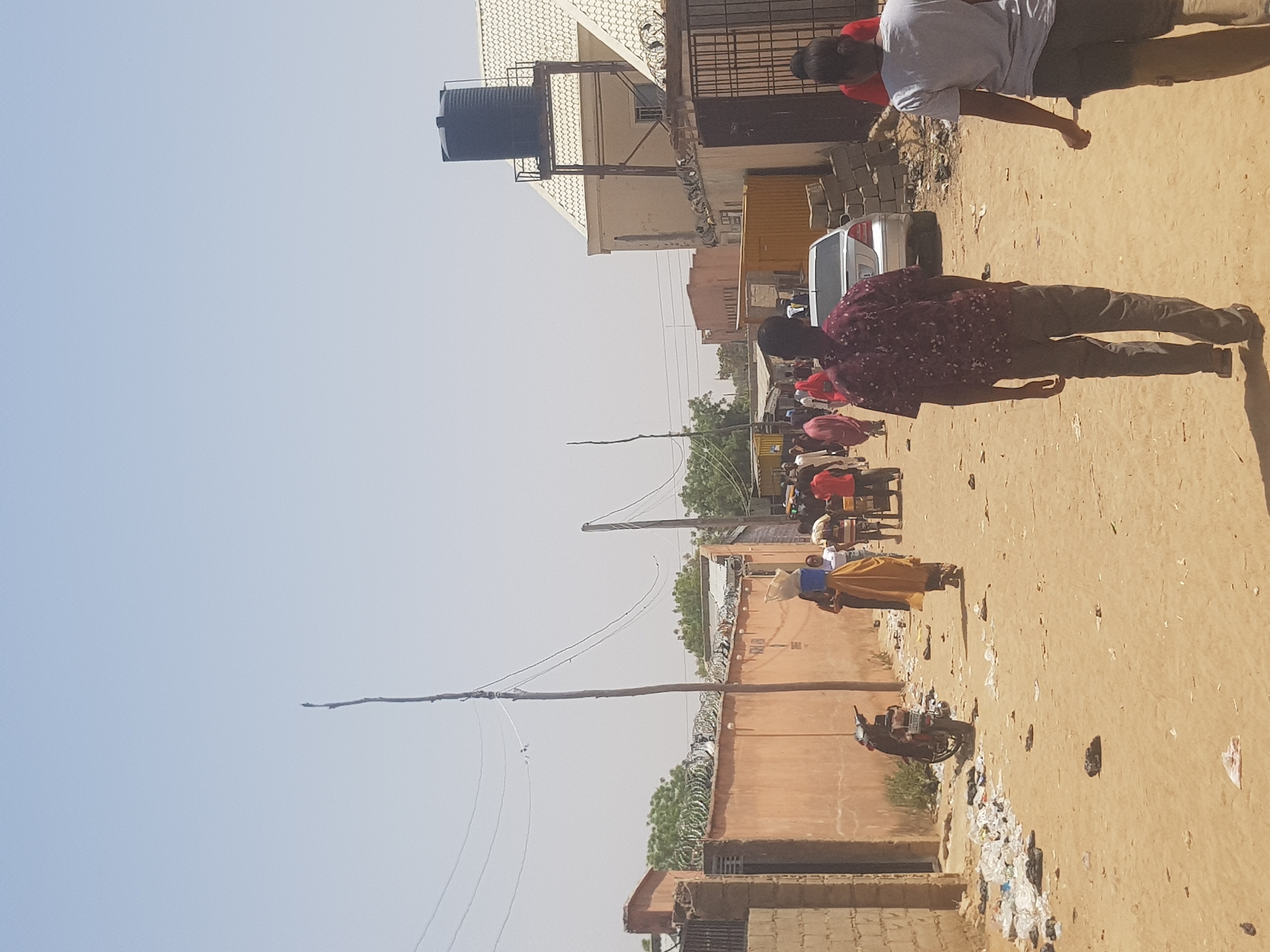 The image captures a street scene in Dabawa, Nigeria. Several individuals are present, including a man in a maroon patterned shirt and gray pants walking on the right side of the frame, and another individual in a light-colored shirt standing near a vehicle. In the background, a group of people appear to be gathered, some possibly carrying items. The environment consists of buildings with barbed wire fencing and overhead utility lines. There is a water tank atop one of the buildings. A motorcycle is parked near a wall. The ground is sandy, and there is visible trash scattered along the street.