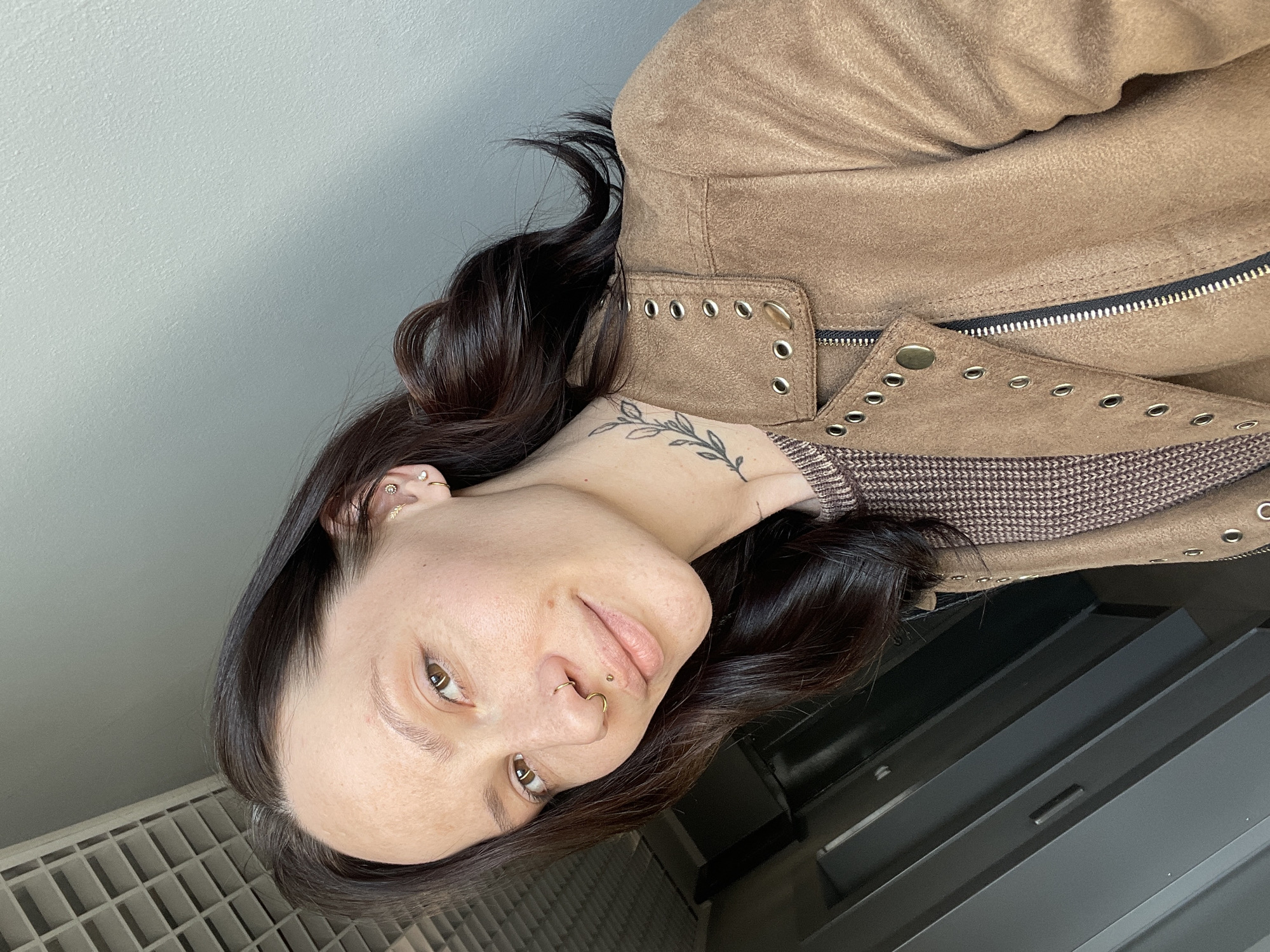 A woman is seen from a close-up angle, positioned at a slant. She has dark brown hair styled with loose waves, brown eyes, and fair skin. Piercings are visible in her ear and nose. She is wearing a brown suede jacket over a brown and white patterned shirt. A tattoo of a leafy branch is visible on her neck. The background includes a light grey wall and a radiator. There is no information available to place the image in the city of Izhevsk, Russia.