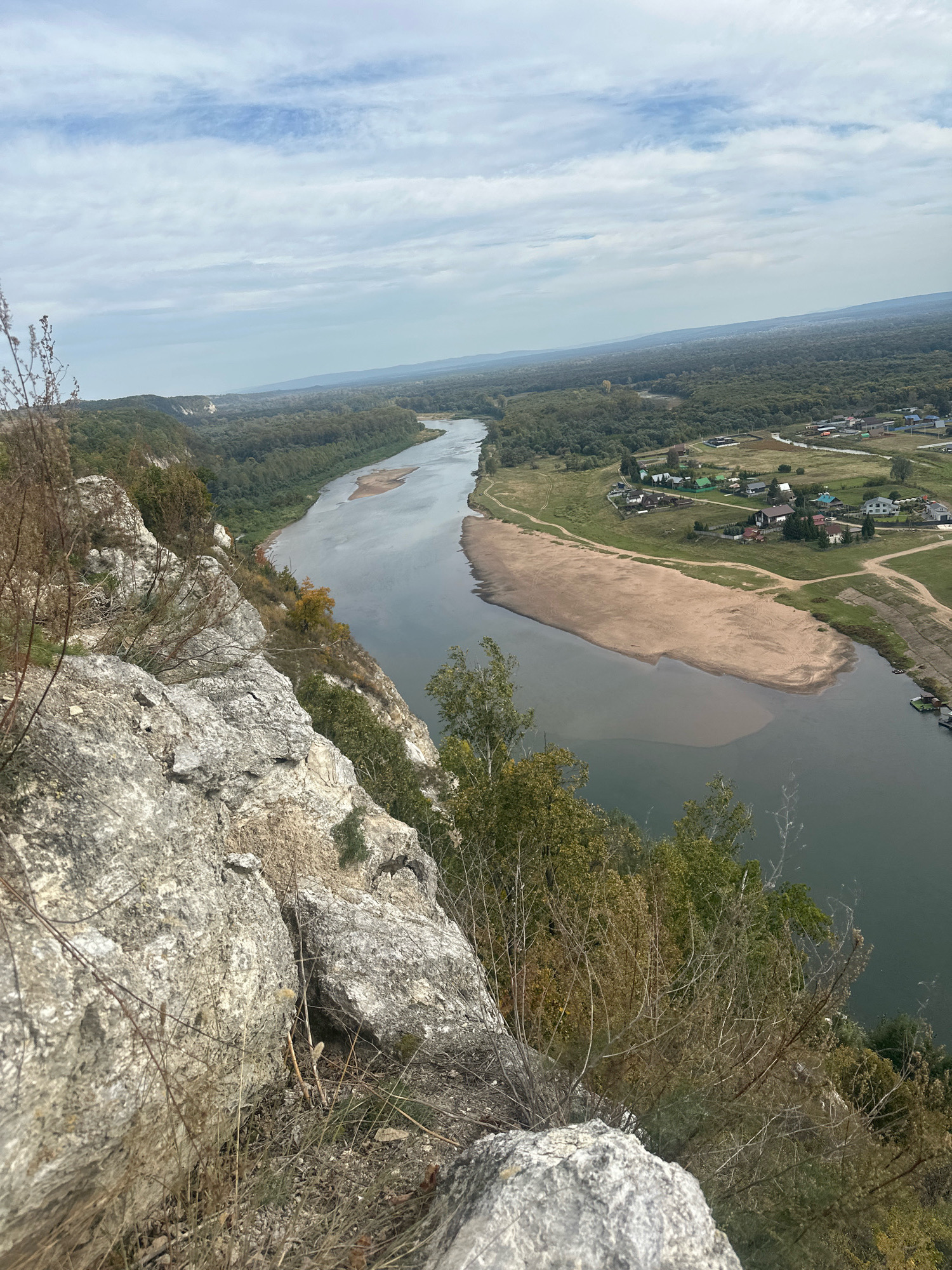 The image is an aerial view from a high vantage point, likely a cliff, overlooking the Sim River in Simskii, Russia. In the foreground, large, weathered grey rocks dominate the left side of the frame, with dry, wispy vegetation growing among them. The river flows from the upper left to the lower right of the image, a wide expanse of grey-green water. A prominent sandbar juts out into the river on the right side of the frame. Across the river, a densely wooded area lines the banks, transitioning into a more open landscape with scattered houses and small buildings, indicating a settlement. The sky above is partly cloudy with a mix of white and grey clouds against a pale blue backdrop. The overall scene depicts a natural landscape with a river and forest, bordering a small rural community.
