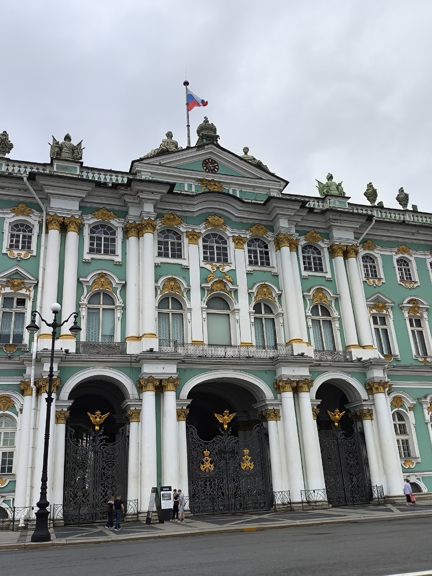 The image shows a frontal view of the Winter Palace in Saint Petersburg, Russia, under an overcast sky. The building is characterized by its striking teal facade, ornate white columns with golden capitals, and intricate gold detailing around the windows and arched entrances. Three large, black wrought-iron gates with prominent golden double-headed eagles and crowns are visible at the ground level. A black lamppost with two lights stands to the left of the main entrance.
In the foreground, on the sidewalk in front of the gates, are four individuals. Two men are walking towards the gates on the left, and two individuals, possibly a man and a woman, are standing near an information board on the right. Further to the right, a woman in a light-colored jacket is walking away from the building, carrying a red bag.
The architecture of the Winter Palace is a prime example of Baroque style, featuring a grand scale and lavish decoration. The building's facade is punctuated by numerous windows, many of which are arched, and crowned with decorative pediments. Statues and sculptural elements are visible on the roofline, and a Russian flag flies from a pole atop the central section of the building. The street in front of the palace appears to be paved with asphalt.