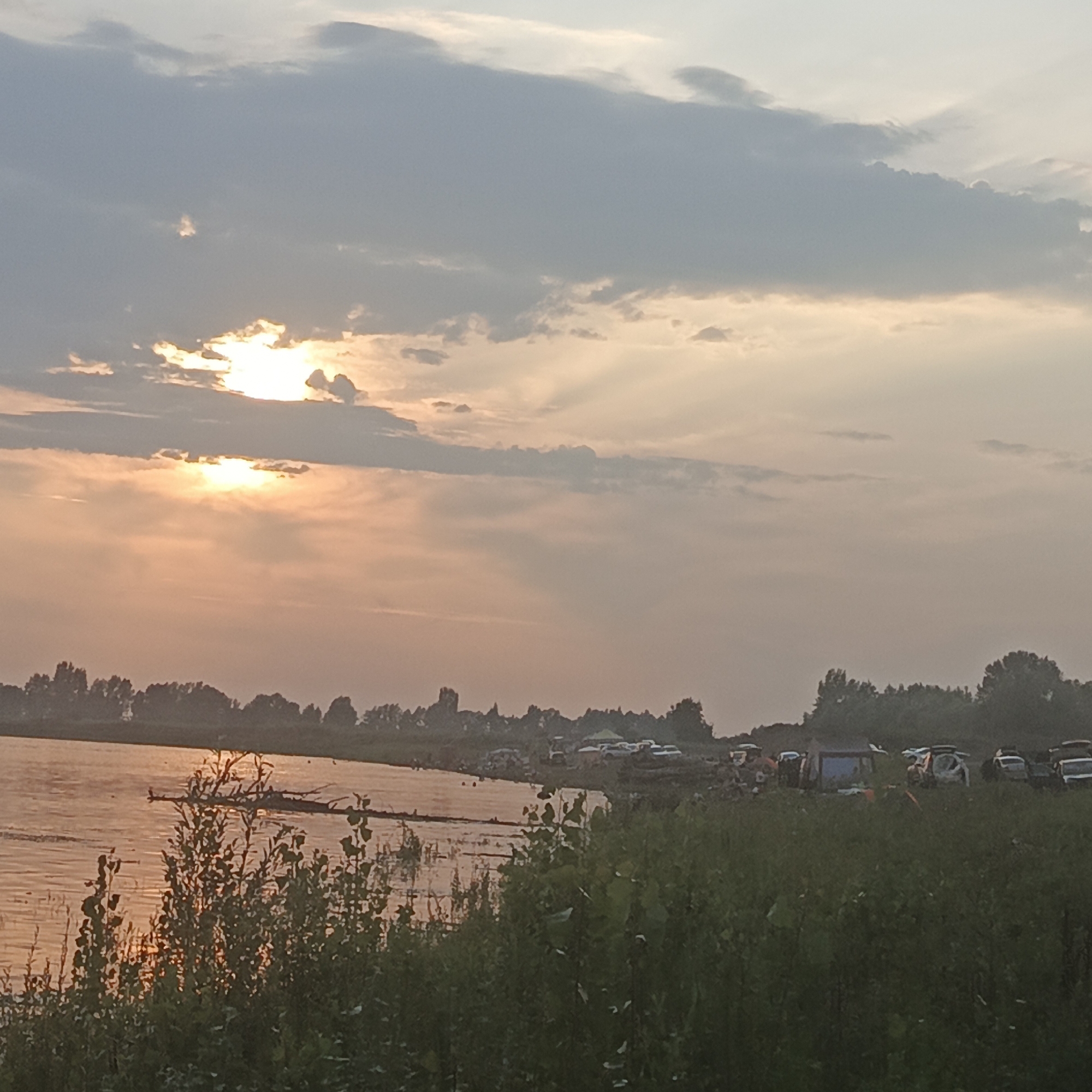 The image depicts a rural landscape at sunset. A body of water, possibly a river or lake, occupies the lower left portion of the frame, reflecting the warm hues of the setting sun. The sun itself is partially obscured by clouds, casting rays of light through the atmosphere. In the mid-ground, a line of trees forms a silhouette against the sky, with a cluster of vehicles, including cars and tents, visible among them. This suggests a gathering or campsite. The foreground is dominated by tall green grass and foliage, obscuring the ground level. The overall impression is one of a serene, end-of-day scene in a natural setting, with evidence of human activity present. The location provided, Старый Буртюк, Russia, indicates a specific geographic context for this rural landscape.