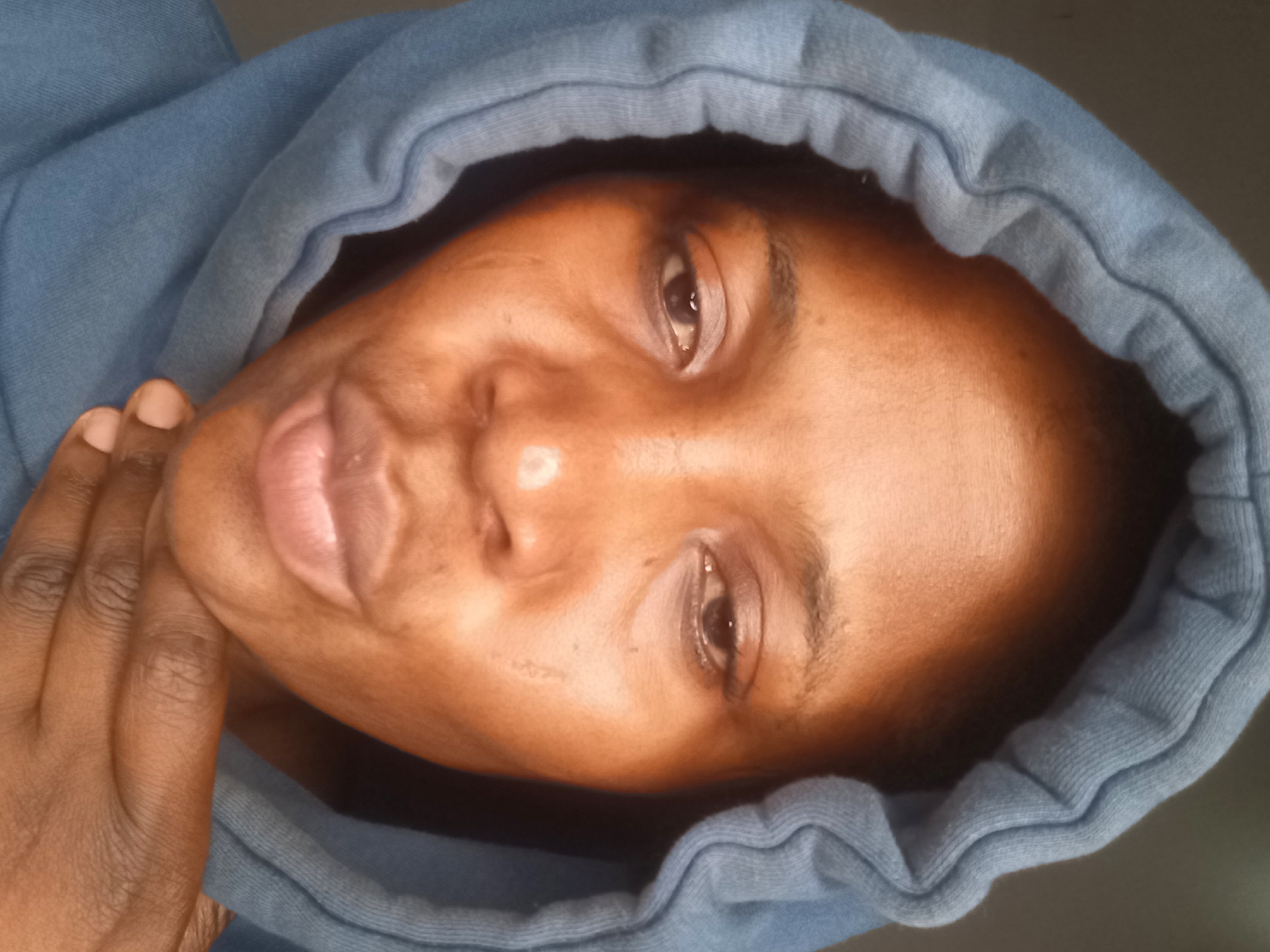 This is a close-up, head-and-shoulders selfie of a Black woman wearing a blue hoodie, with the hood pulled up to frame her face. She is looking downwards with a neutral or slightly somber expression. Her hand is resting on her chin. The background is a plain, dark, out-of-focus surface, suggesting an indoor setting. The lighting is from above and slightly to the front, highlighting her features and creating some sheen on her skin. There are no identifiable landmarks or context to suggest a specific location within Jos, Nigeria.