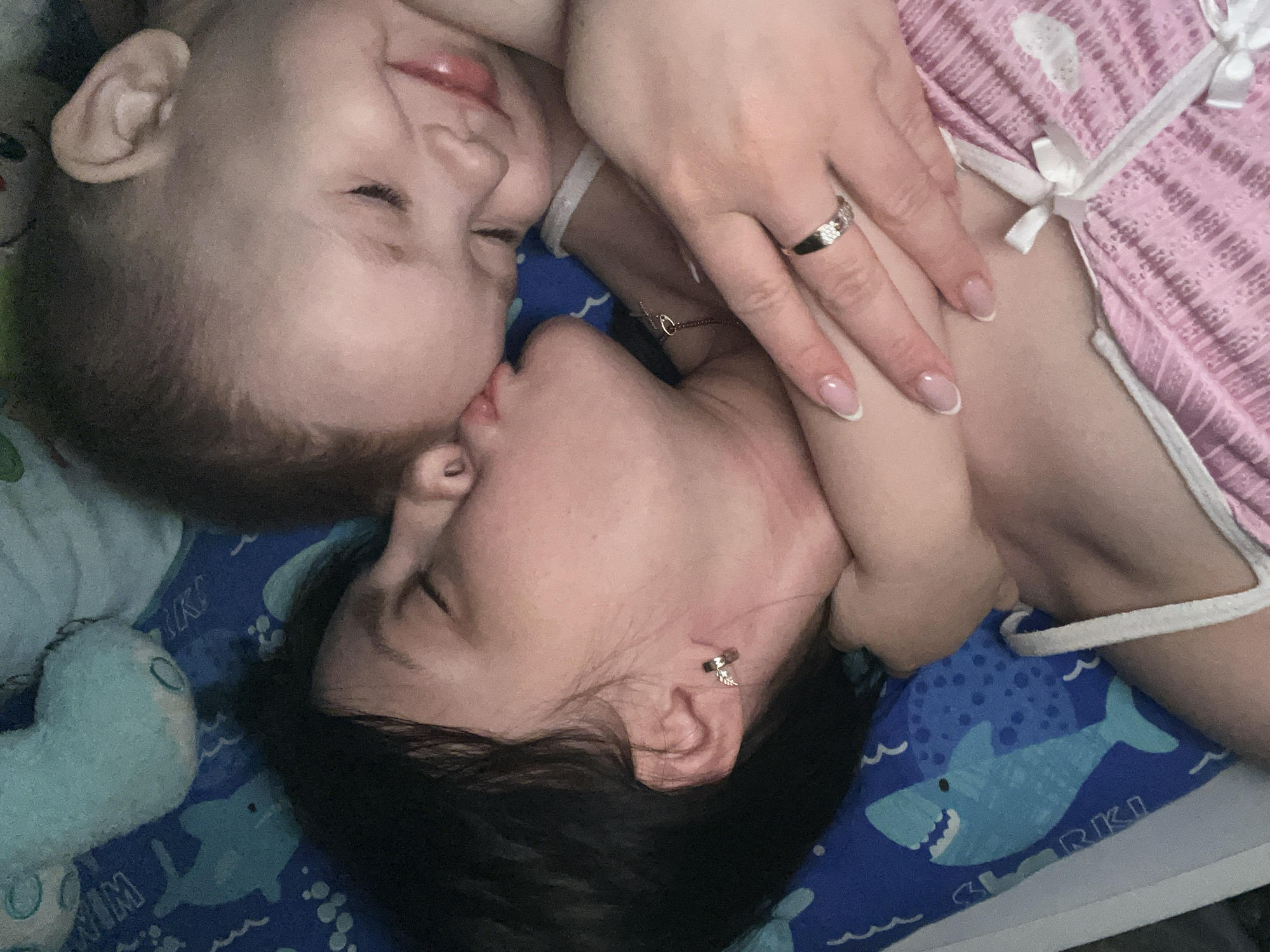 A woman with dark hair is kissing a young child on the lips. The woman is wearing a ring on her left hand and an earring in her left ear. The child has fair skin and light-colored hair. They are lying on a blue and white patterned blanket with a stuffed animal nearby. The blanket features shark graphics and the word "SHARKI" on it. The image appears to be taken indoors. The location in Марковский, Russia is not confirmed, as there are no identifiable landmarks or clues in the image to verify it.