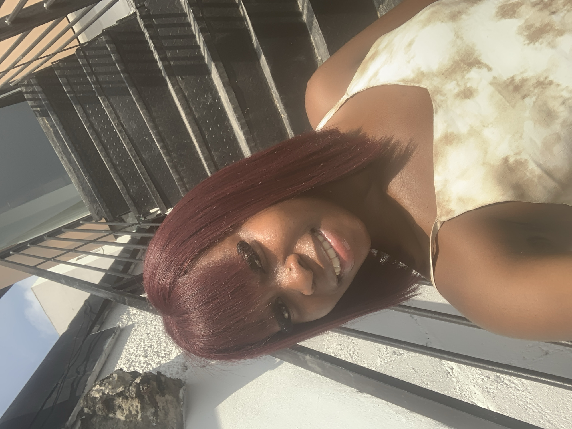 A dark-skinned woman with burgundy shoulder-length hair and bangs smiles at the camera. She is wearing a light-colored dress with a brown tie-dye pattern. She is standing on a metal staircase outdoors, with a white textured wall to her left. The sun is shining, casting shadows on the wall and stairs. The location is the city of Moba, Nigeria.