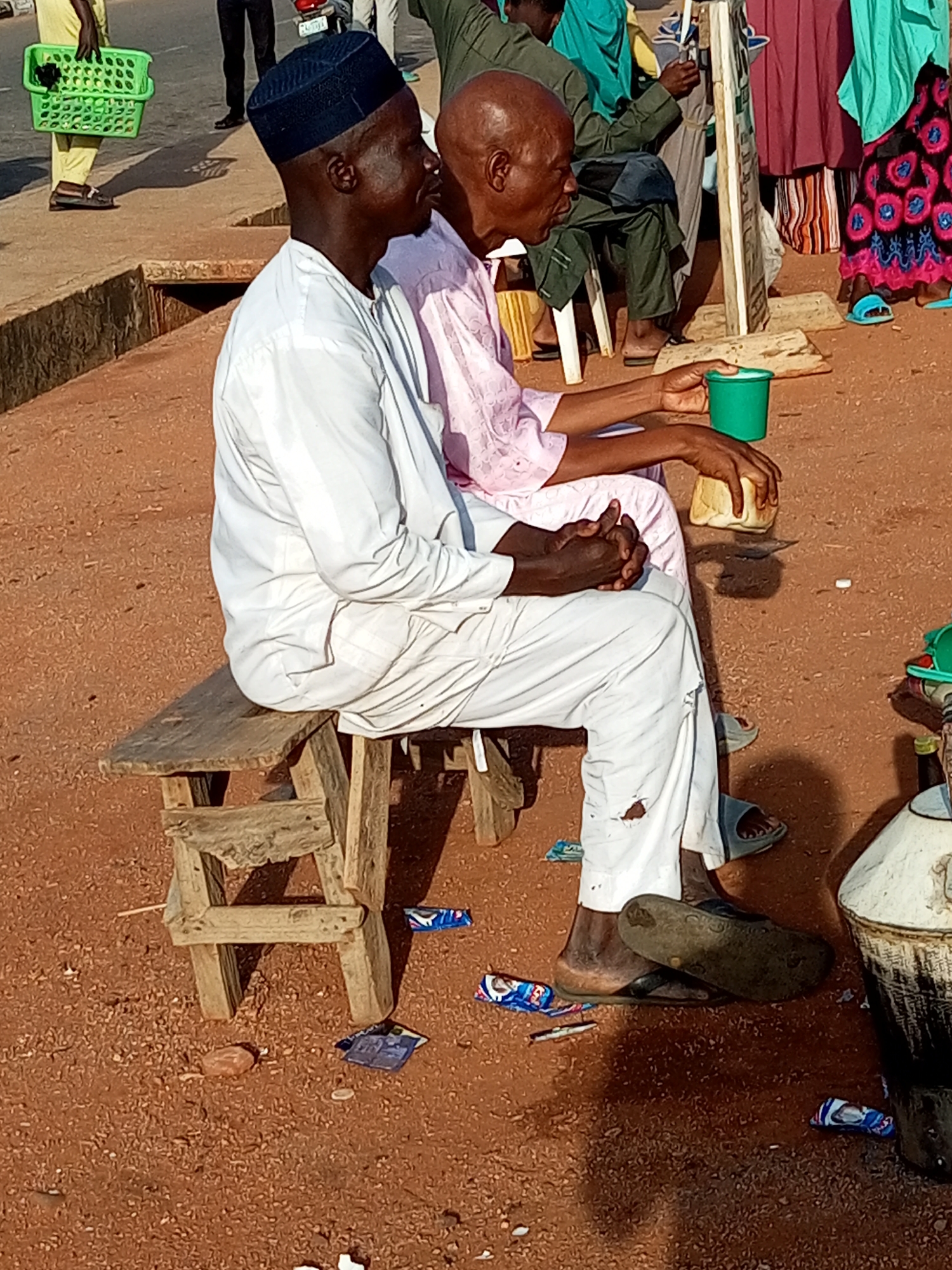 In a city location of Unknown, Unknown, two men are seated on a rustic wooden bench. The man on the left is wearing a white traditional outfit and a black cap. He has his hands clasped in his lap. The man on the right is wearing a light pink traditional outfit. He is holding a piece of bread in his left hand and has a green cup to his right. The ground is covered in reddish-brown dirt and scattered with debris, including wrappers and a small white object. Behind the men, other individuals are visible, some wearing colorful clothing, and a green basket is being carried by someone in the background. A white sign is propped up on wooden supports near the man on the right. The scene appears to be outdoors, possibly in a market or public waiting area.