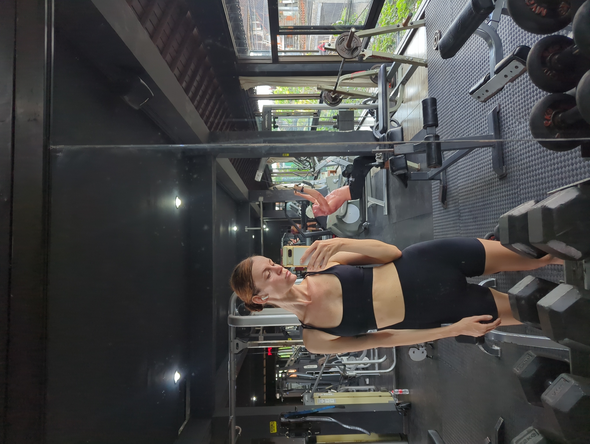 The image shows a woman in a black sports bra and shorts holding a smartphone, appearing to take a selfie in a gym. The gym, likely located in Mengwi, Indonesia, has various exercise machines and equipment. A person is seen using a machine in the background. The area is well-lit, and the environment appears to be a standard fitness center.