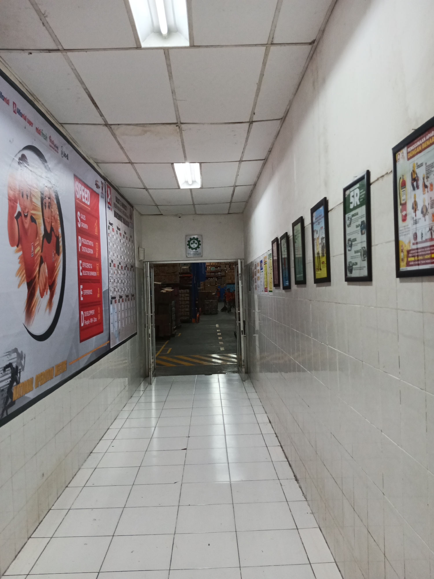 The image depicts an interior hallway, likely within a warehouse or factory, featuring a long, tiled corridor. The hallway is illuminated by fluorescent ceiling lights and is lined with white tiled walls. On the left wall, a large banner displays animated characters and text in red and white. Further down the left wall, a series of smaller posters or charts are visible. The right wall is adorned with several framed pictures or posters, each with distinct graphics and text. At the far end of the hallway, a doorway leads to a storage area filled with boxes and shelving. A safety sign with a green circular logo and text is also mounted on the wall near the doorway. The floor is covered in white tiles with visible grout lines, and a yellow and black striped hazard marking is present near the entrance to the storage area. The overall environment appears functional and industrial. No specific location in Makassar, Indonesia is evident from the visual information provided.