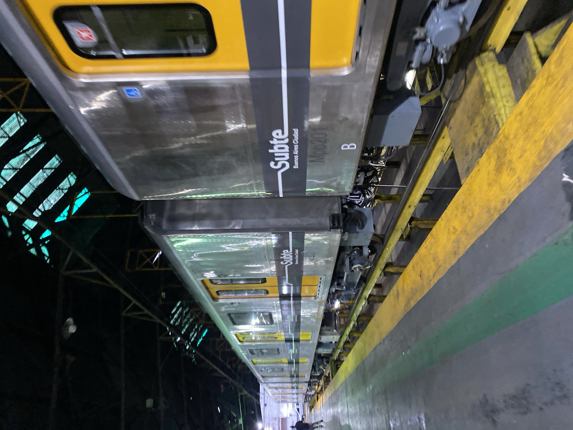 The image depicts a train of the Buenos Aires Metro (Subte) in what appears to be a maintenance facility or depot in Buenos Aires, Argentina. The train cars are painted with a silver or metallic finish and marked with a yellow stripe near the top, and a vertical dark gray stripe with the word "Subte" and "Buenos Aires Ciudad" written on it. The train's number is "Mpb201" and car letter is "B". The train is situated along a yellow painted platform edge. The warehouse structure is visible in the background, with metal supports and some bluish lighting. There is a person standing near the end of the train.
