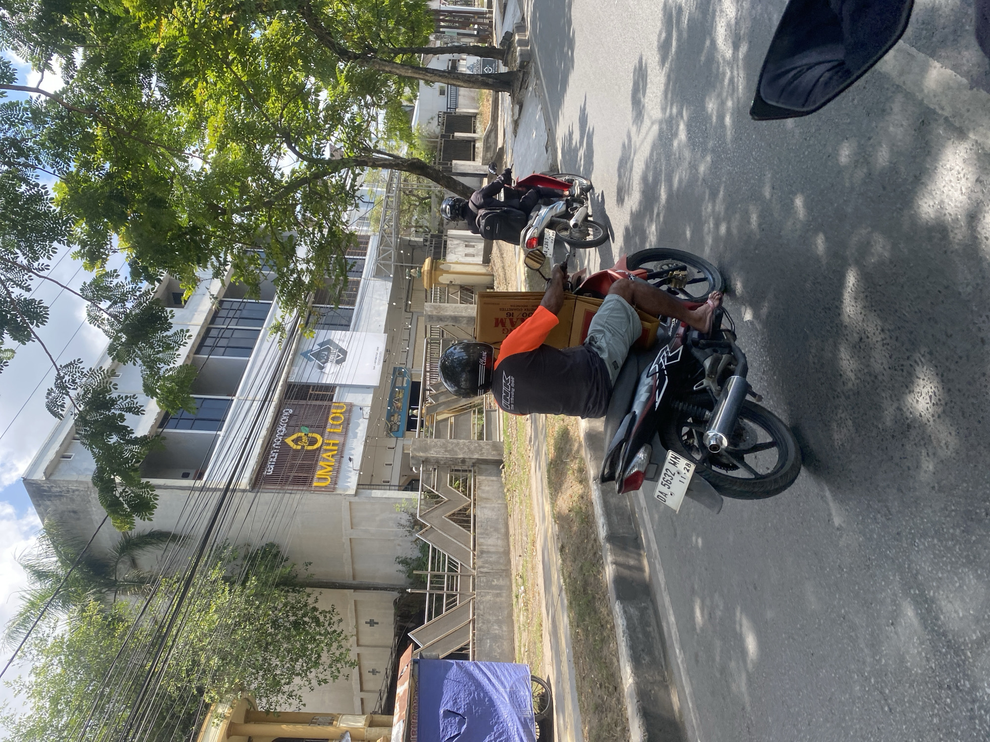 A side-view, eye-level shot captures a street scene in Banjarmasin, Indonesia, on a sunny day. The foreground shows the rear portion of a motorcycle, with its black license plate reading "DA 5632 MN 11-28" visible. To the left, another motorcycle is partially visible, carrying a driver and a large cardboard box behind them. The driver of this motorcycle wears an orange and black shirt, light-colored shorts, and flip-flops.
The background features a multi-story building with a light-colored facade and a prominent sign with yellow lettering reading "UMAH LOU" and "teman nongkrong". Trees with lush green foliage line the street, casting dappled shadows on the asphalt. A blue tarp-covered structure on wheels is parked on the side of the road near the building. The scene conveys a typical urban environment in Indonesia, with everyday life unfolding on the street.