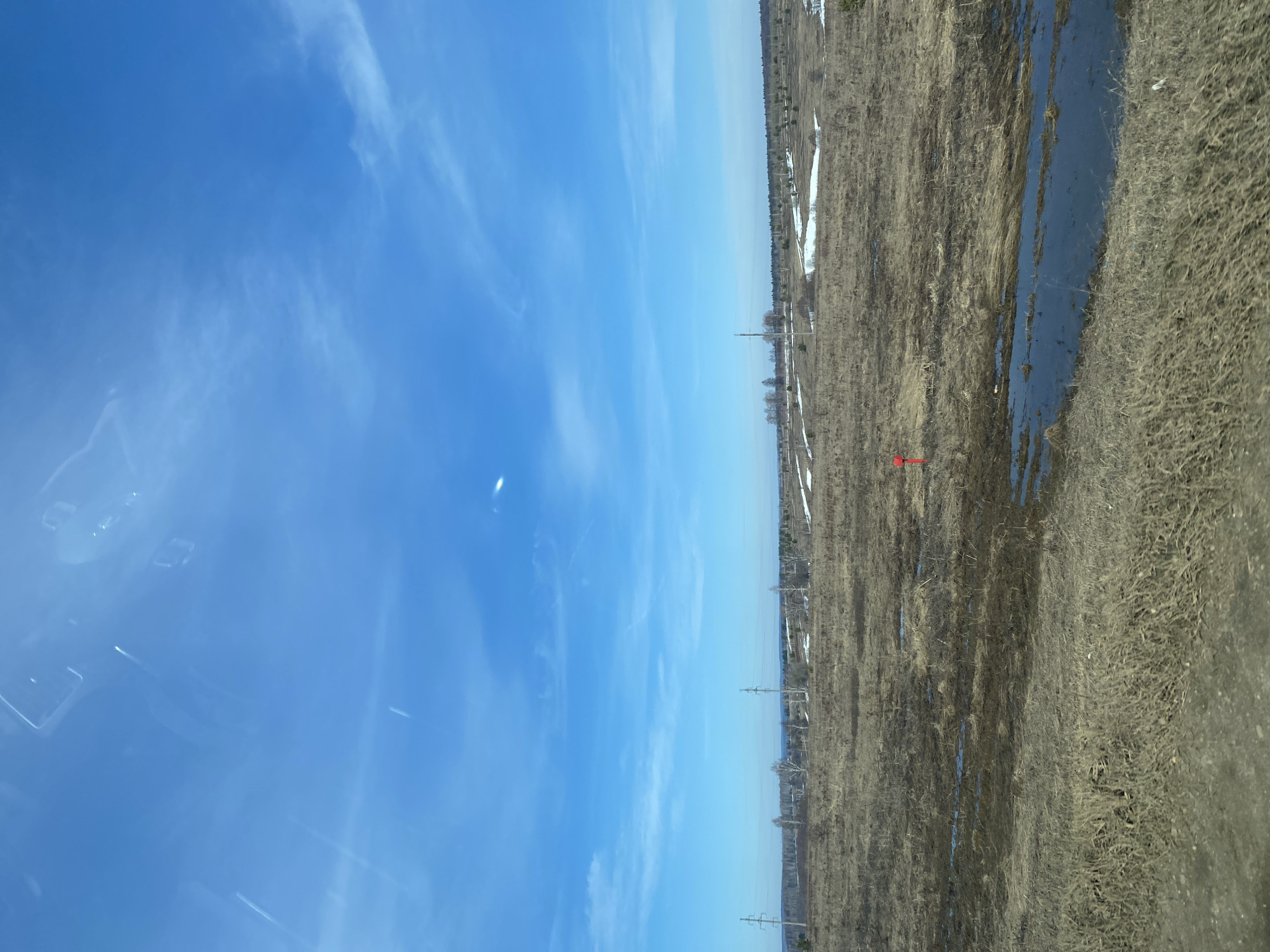 The image shows a landscape in Kueda, Russia. The sky is a clear blue with scattered cirrus clouds. Below the sky is a rural landscape with a field partially covered in dried vegetation and patches of melting snow. Power lines stretch across the horizon. A narrow stream or standing water runs through the field. A red arrow is superimposed on the image in the center of the field.
