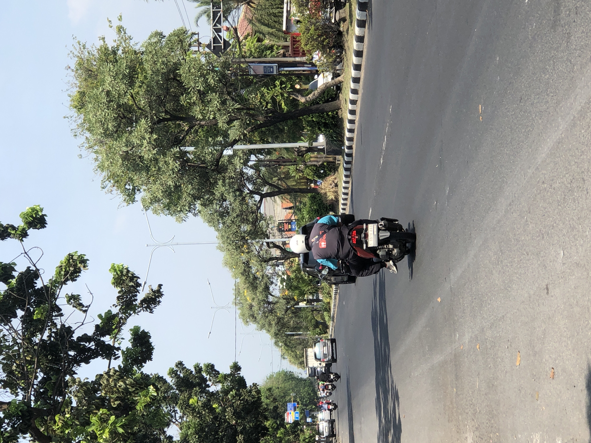 In Larangan, Indonesia, on a sunny day with clear skies, a motorcycle carries two individuals. The rider, wearing a white helmet, is dressed in a dark blue jacket with a visible logo featuring a red and white emblem and the word "You*ube" in white. The passenger, seated behind the rider, wears a dark jacket and jeans. The motorcycle, a red and black model, displays a license plate that reads "S 6978 DAD" and has a white rear fender. They are on a paved road with two lanes in the direction of travel. The road is bordered by a black and white striped curb. On the right side of the road, there are trees with lush green foliage, a sidewalk, and a few parked cars, including a white car. On the left side, more trees and a traffic light can be seen. Several other motorcycles and cars are visible further down the road, suggesting moderate traffic. The scene appears to be an urban or suburban street.