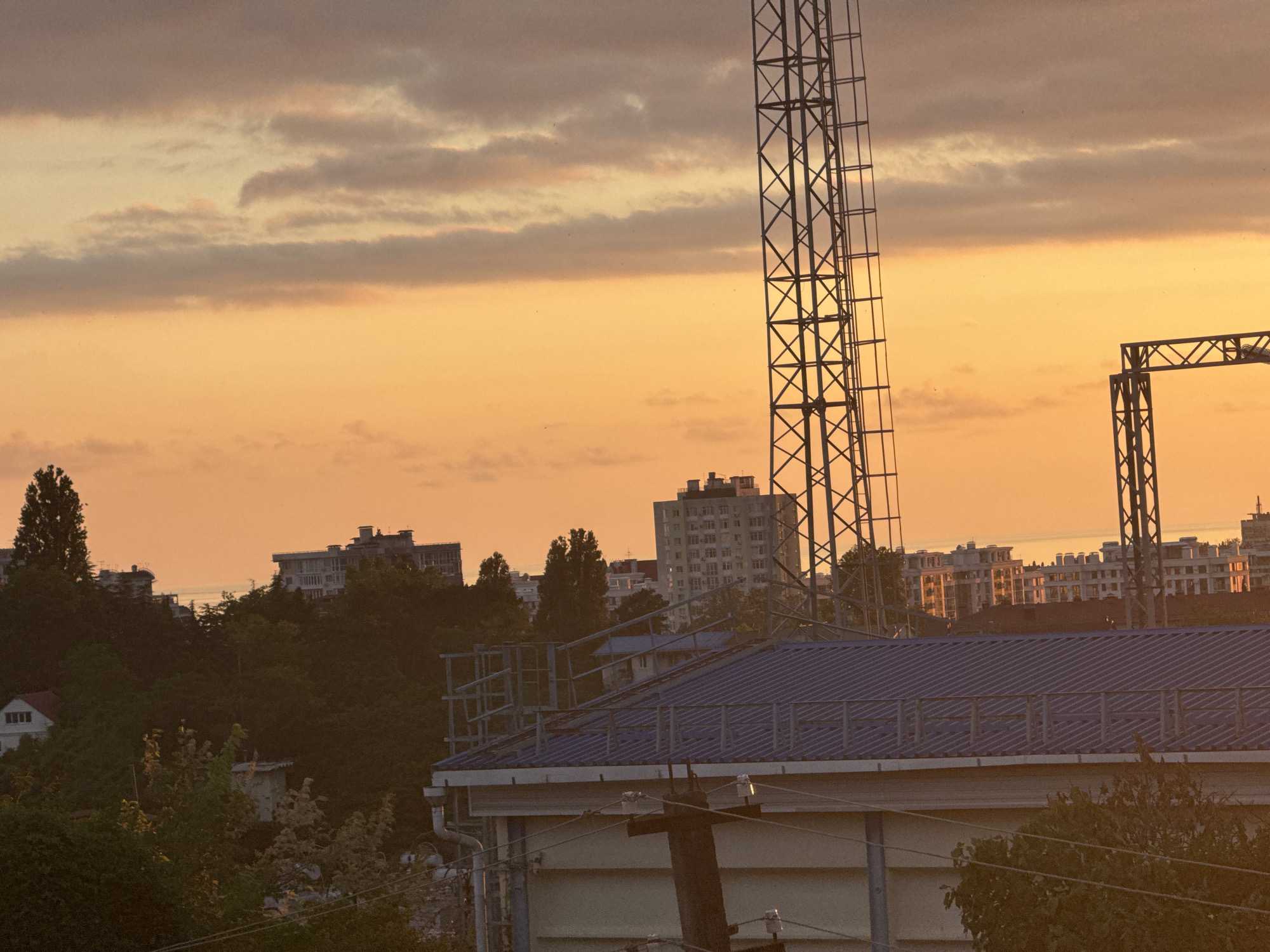 The image depicts an urban landscape at sunset, likely in Sochi, Russia. The sky is a warm gradient of orange and yellow, with soft, wispy clouds scattered across it, indicating fair weather. Dominating the foreground is the blue, corrugated metal roof of a building, with a visible gutter system and overhead electrical wires. Further back, a dense canopy of dark green trees covers a hillside, punctuated by several apartment buildings with white facades and numerous windows. Tall, lattice-like metal structures, possibly part of a telecommunications tower or industrial equipment, rise prominently against the sky on the right side of the frame. The overall impression is one of a bustling city transitioning into evening, with a blend of natural and man-made elements. The lighting suggests it is late afternoon or early evening, with the setting sun casting a warm glow over the scene. No people are visible. There is no discernible text in the image.