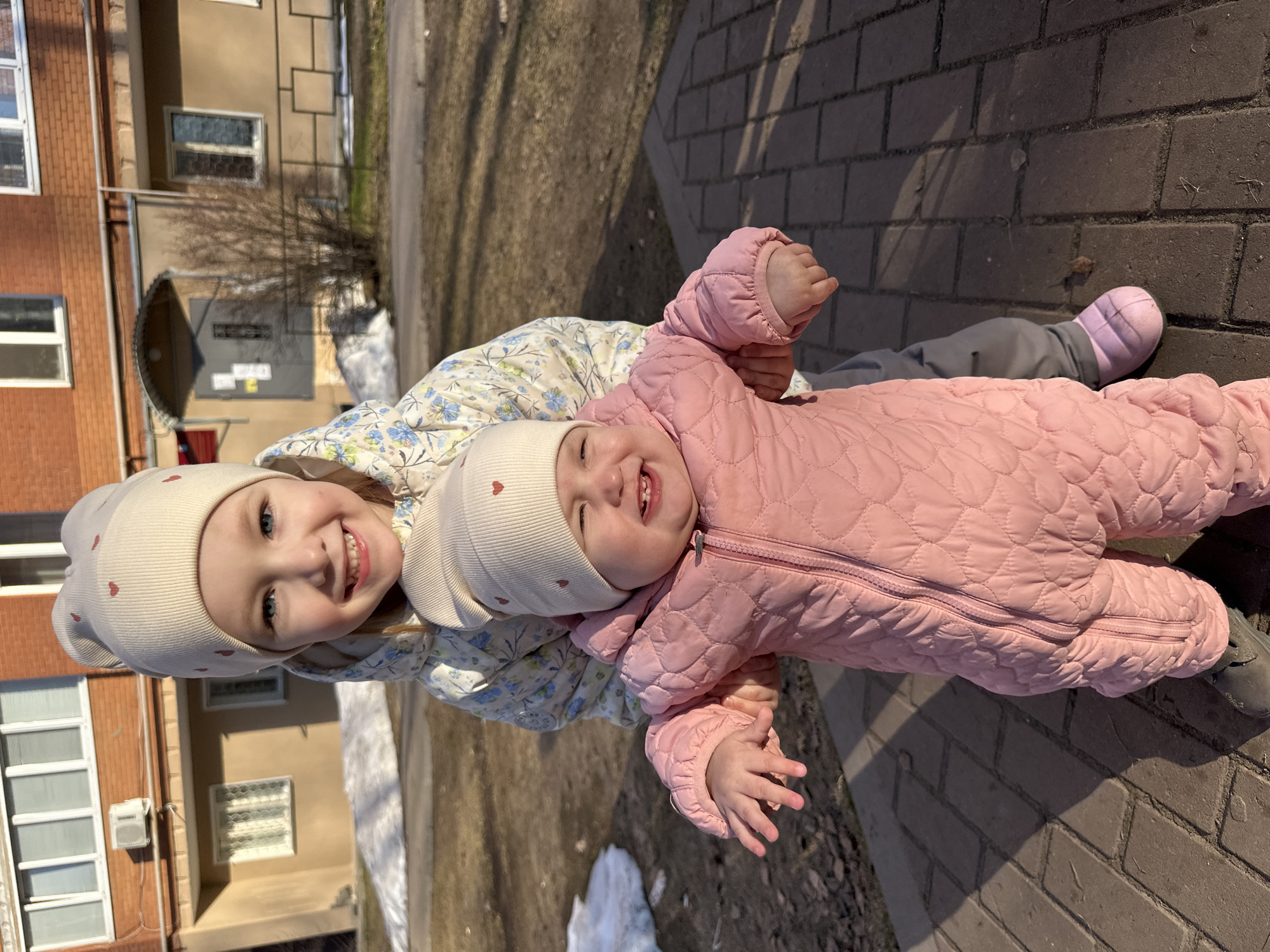 Two young girls are the primary subjects of the image. The older girl is standing and holding a younger girl, who is lying horizontally in her arms. Both girls are wearing matching knit hats with a small heart emblem. The older girl is also wearing a light-colored floral jacket, while the younger girl is wearing a pink quilted snowsuit.
The setting appears to be an outdoor public space, possibly a park or plaza, in the city of Chaikovsky, Russia. This inference is based on the urban architecture visible in the background, which includes multi-story buildings with windows, brick facades, and external features consistent with typical Russian urban design. The ground is paved with interlocking bricks, and there are remnants of snow in the vicinity.
The image captures a moment of interaction between the two girls, with the older girl smiling and the younger girl appearing to laugh. The scene is well-lit, suggesting it is daytime.