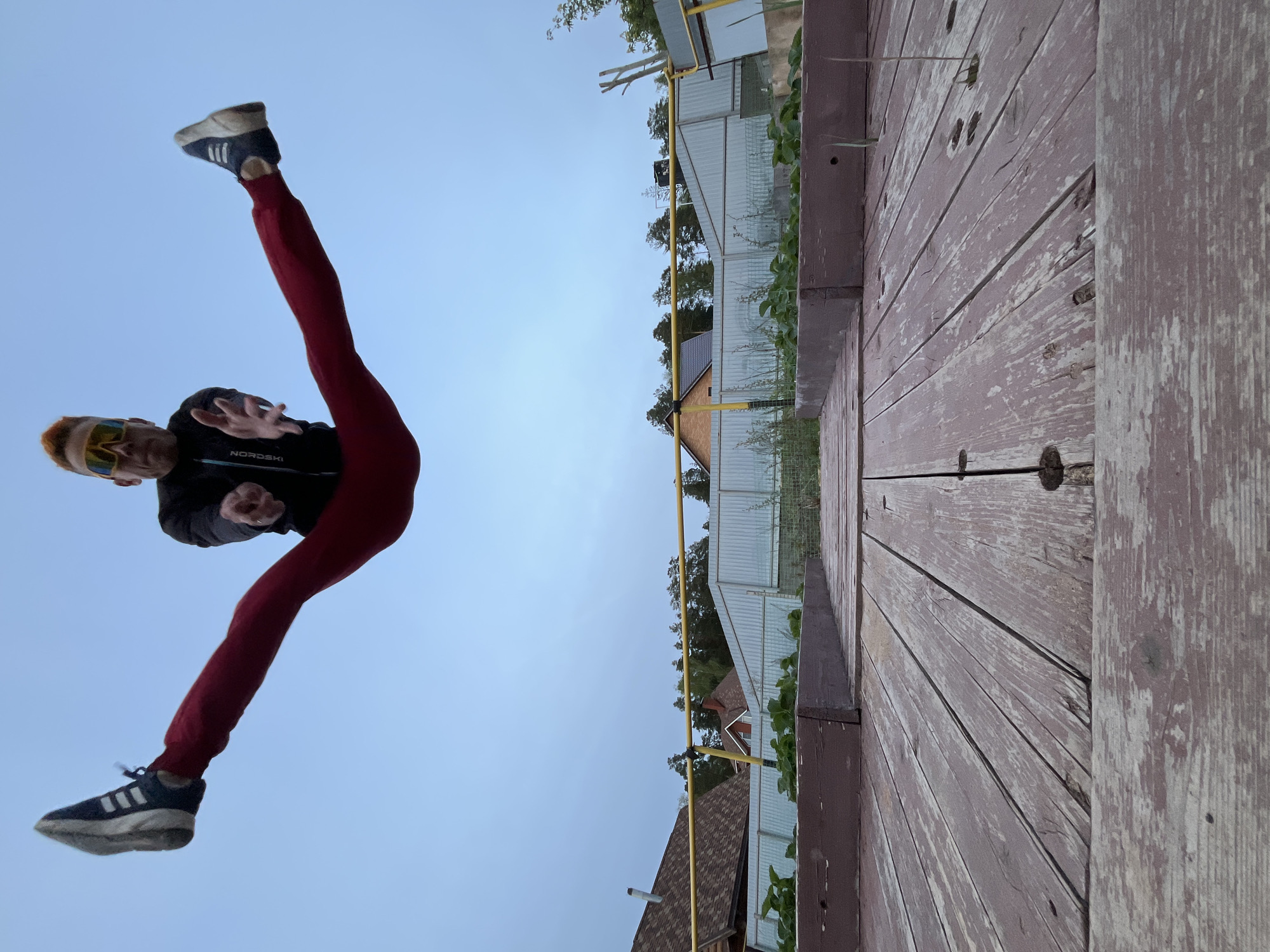 A person is suspended in mid-air against a backdrop of a pale sky, with one leg extended upwards and the other downwards in a straddled position. The person is wearing a dark jacket, red leggings, and dark sneakers with white stripes. They appear to be wearing sunglasses.
The background includes elements of a residential area. There is a wooden structure or deck in the foreground with peeling paint. Behind the deck is a partially visible fence with metal mesh and supporting yellow posts. Beyond the fence, rooftops of houses are visible, interspersed with trees.
The image was taken in Chaikovsky, Russia.