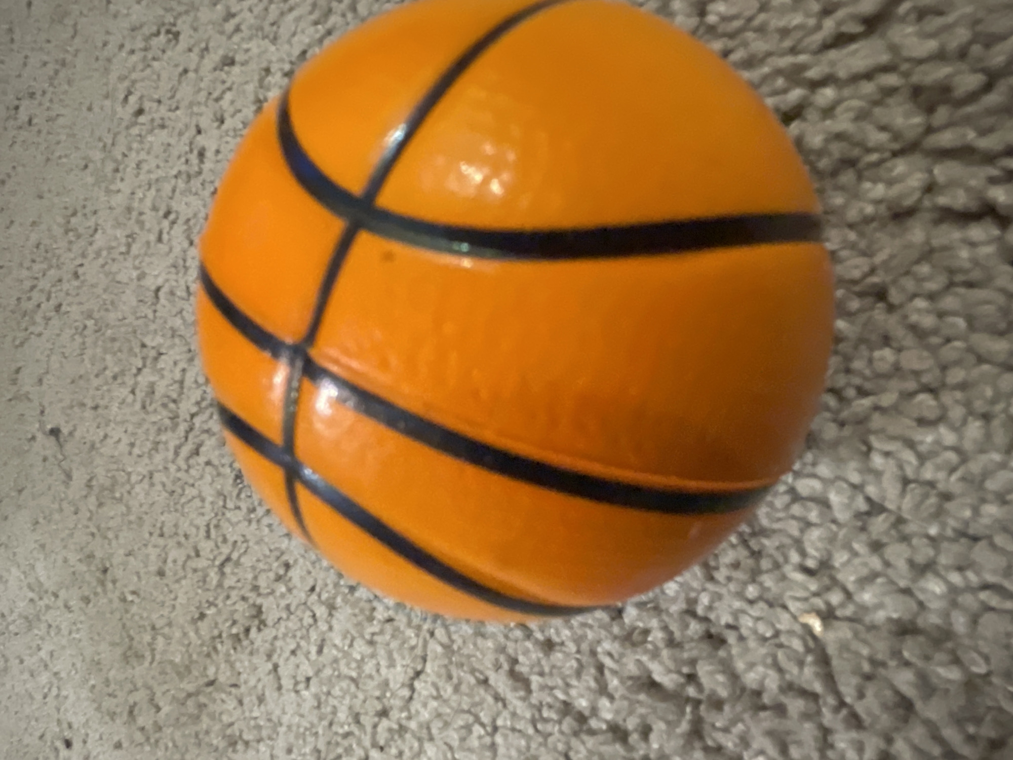An orange basketball with black lines sits on a grey carpet. The basketball is positioned slightly off-center to the right. The carpet appears to be a standard indoor carpet with a textured surface. The lighting is overhead, casting subtle shadows. There are no people or other objects visible in the image. The scene does not provide any specific details that would allow for localization within Tallinn, Estonia.