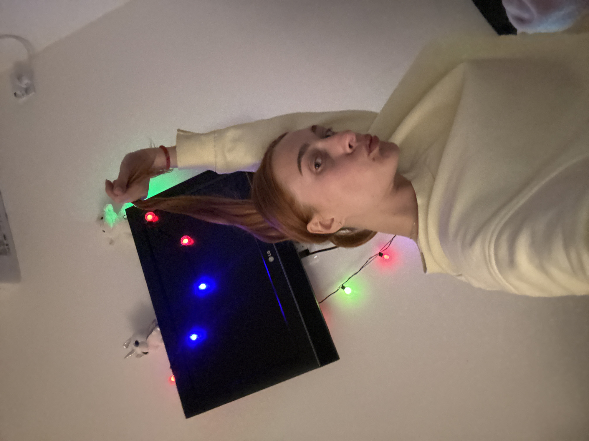 The image presents a person lying near a television screen illuminated with blue light, surrounded by a string of colorful Christmas lights. The individual, presumably in an indoor setting, is wearing a light-colored sweatshirt. A white wall with an electrical outlet is also visible. The location, Budva, Montenegro, is not verifiable from the given image.