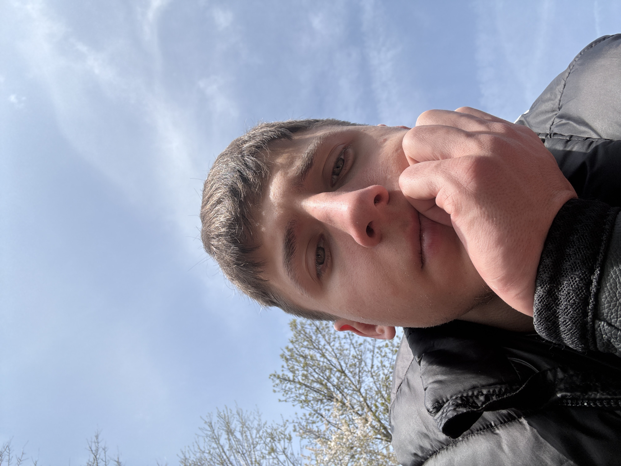 A low-angle close-up shot shows a young man with short brown hair, fair skin, and visible stubble. He is wearing a black puffy jacket. His right hand is near his face, fingers slightly curled. The background features a blue sky with white clouds and the bare branches of a tree with budding white flowers visible near the bottom of the frame. The location appears to be outdoors during the day, potentially in or near the city of Bozhurishte, Bulgaria.