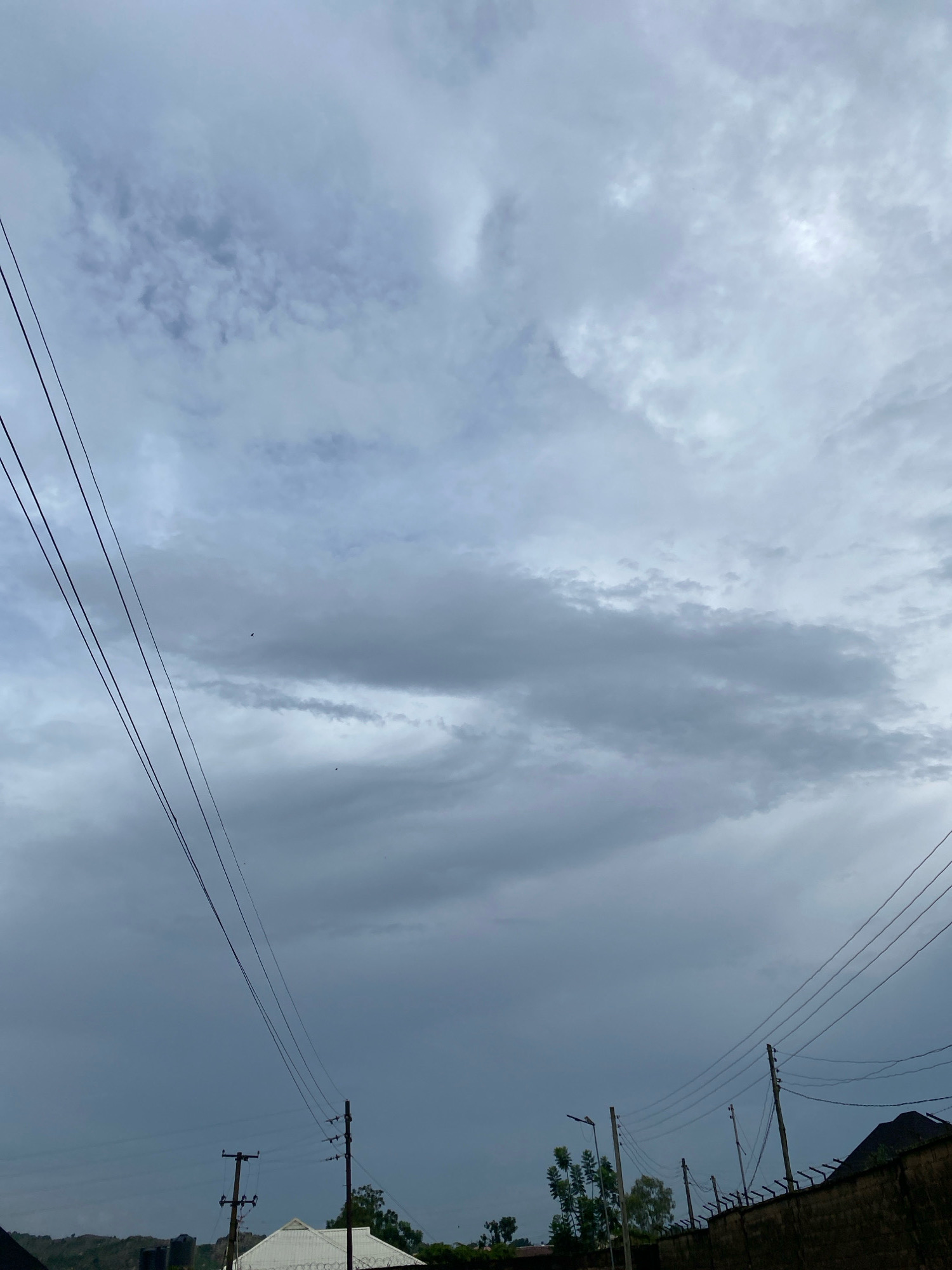 The image depicts an overcast sky above a somewhat built-up area. Three dark power lines stretch diagonally across the frame from the upper left to the lower right. In the lower portion of the image, several utility poles with insulators and wires are visible, along with parts of buildings and a textured wall topped with barbed wire. A few trees are scattered among the structures. The sky is filled with gray, textured clouds, suggesting a cloudy or potentially rainy day. The scene appears to be an outdoor urban or semi-urban environment. The specific location is identified as Jos, Nigeria.