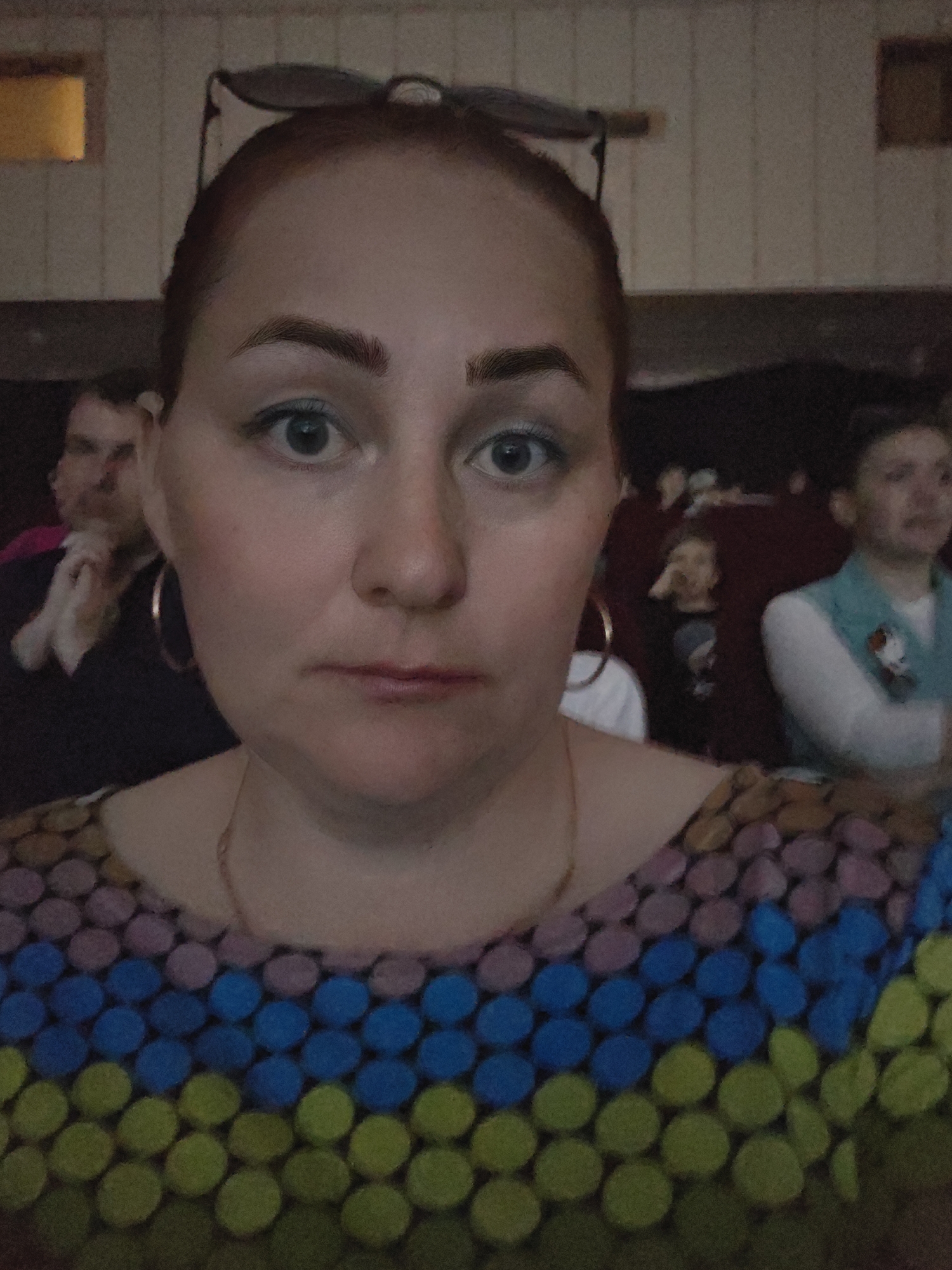 A medium indoor shot depicts a woman in the foreground, with other people sitting behind her in a theater setting, location Unknown, Unknown.
The woman in the foreground is fair-skinned with reddish-brown hair pulled back, large gold hoop earrings, and a thin gold necklace. She wears a multicolored shirt with rows of blue, purple, and green dots. The light is focused on her face and shirt. She has an unsmiling, neutral expression. Her eyebrows are thick and well-defined.
Behind her, other people are seated in what appears to be theater seating. To the left is a man with his hand to his face, looking toward the camera's left. Further to the right and slightly behind the woman, other people are visible, including a young child with their hand near their mouth and a woman in a blue vest.
The background is dark and somewhat blurry, typical of indoor, low-light conditions. A rectangular window is faintly visible in the top left corner of the image. The setting seems to be a performance or event venue. The overall image quality is grainy, indicating a high ISO setting or a lower-quality camera.
