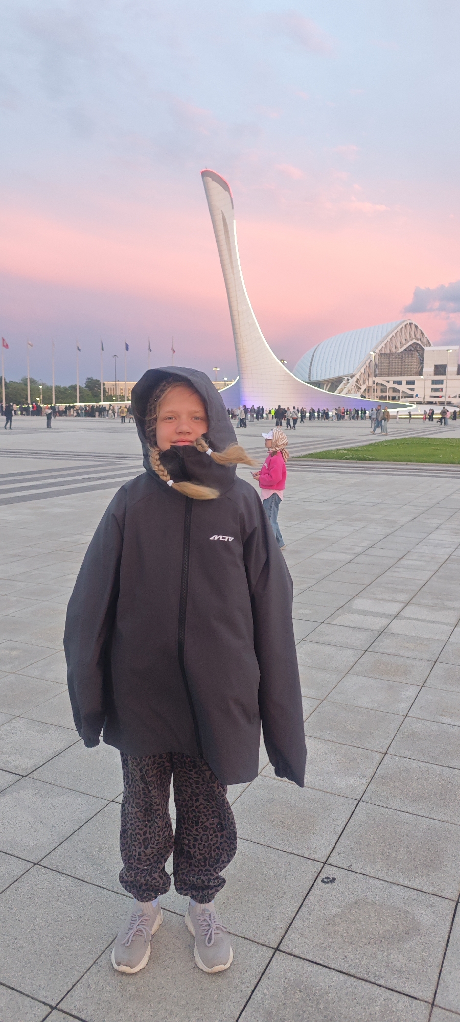 A young girl stands centered in the foreground, wearing a black hooded jacket with the logo "ACTIV" visible on the chest. She has blonde hair styled in braids that protrude from the hood. The jacket is paired with leopard-print trousers and light-colored sneakers.
Behind her, the Sochi Olympic Park in Sirius, Russia, is visible. The Olympic cauldron is a prominent architectural structure in the background, with its distinct torch design reaching towards the sky. In the distance, the Iceberg Skating Palace and other structures from the Olympic complex are seen, along with several flags.
The area is a large, open plaza with a tiled surface. People are walking around, though slightly blurred in the distance. The sky is a mix of pink and blue, suggesting either sunrise or sunset.
