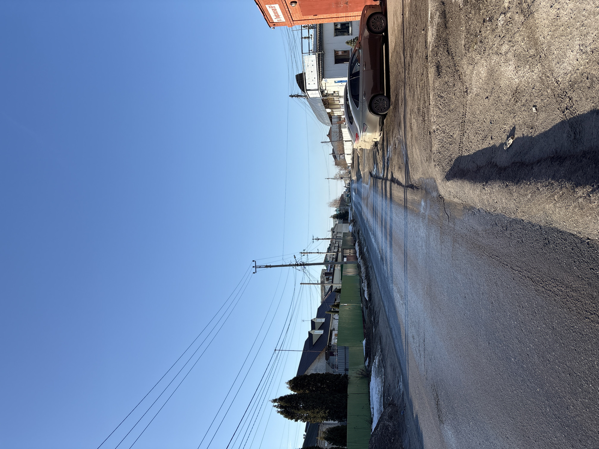 The image depicts an urban street scene at Profsoyuznaya Street 56 in Ulyanovsk, Russia, under a clear, bright blue sky. The asphalt road, running from the foreground into the distance, appears wet and reflective in patches, indicating moisture or ice. Patches of dirty, melting snow are visible along the road edges on both sides.
On the left side of the street, a green corrugated metal fence encloses properties featuring various residential or commercial buildings. Several evergreen trees, including a pair of tall, dark conifers, are situated behind this fence. Overhead, multiple black utility lines traverse the frame, supported by wooden and metal utility poles positioned along the street.
On the right side of the street, a dirt and mud shoulder parallels the road. Two sedan vehicles are parked on this shoulder, facing away from the camera. The foreground vehicle is a light brown or gold sedan, and a silver or grey sedan is partially visible behind it. Adjacent to these vehicles is a red brick building with a large red metal garage door. A white sign affixed to this building clearly displays "ул. Профсоюзная 56" (Profsoyuznaya Street 56). Behind this red structure, a larger white building with an arched roof and a raised platform is visible. Long shadows are cast across the dirt shoulder and parts of the road from unseen objects to the left of the frame. No individuals are visible in the scene.