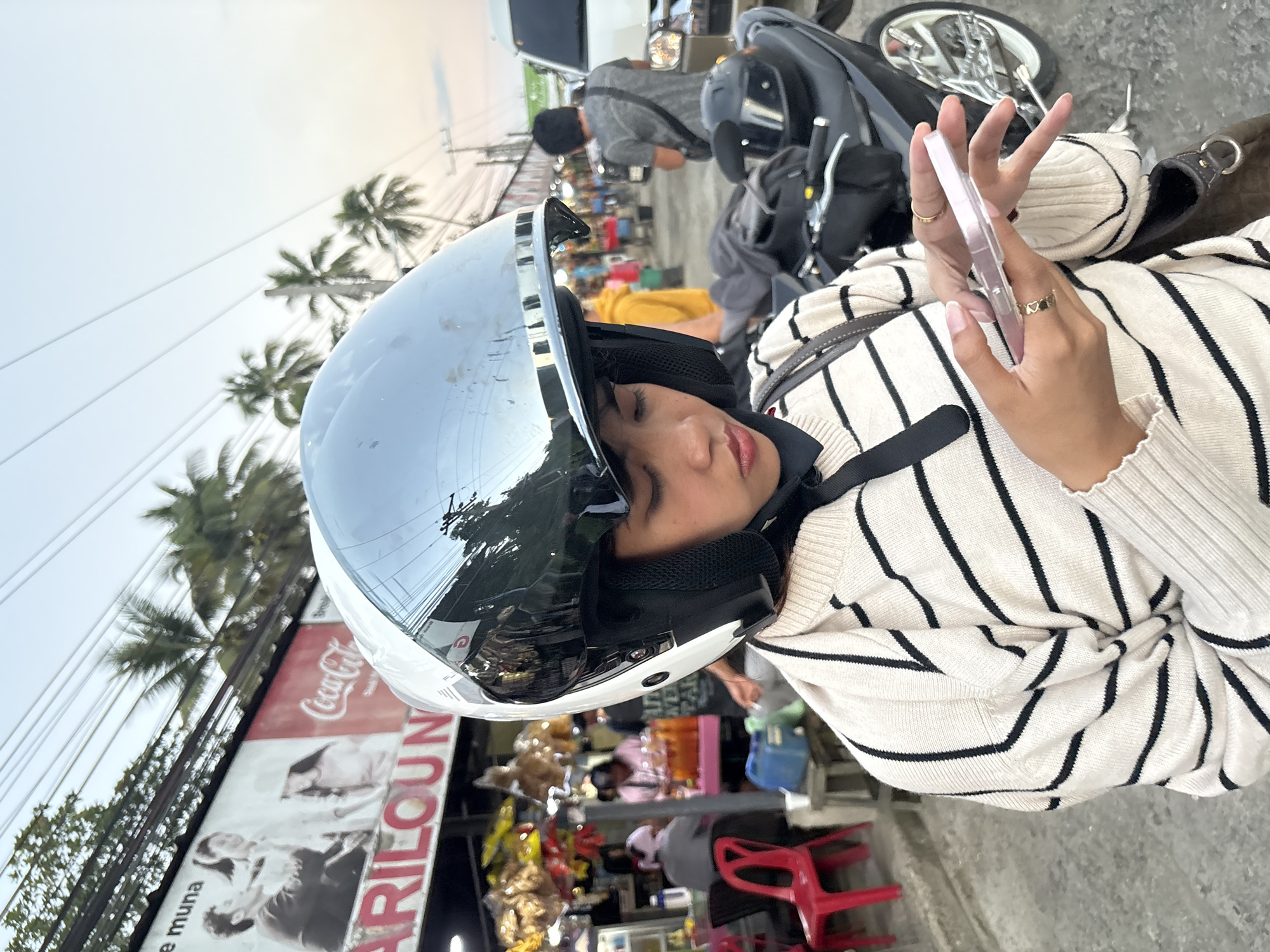 A young person wearing a white and black striped sweater and a motorcycle helmet with a reflective visor is looking down at a pink smartphone in their hands. They are standing on a street with a motorcycle parked behind them. In the background, there are shops and palm trees, suggesting a tropical location. A large billboard for Coca-Cola is visible, along with a sign that reads "ARILOU N." The overall impression is that of a casual street scene in a town or city, possibly during the late afternoon or early evening given the lighting. The location is identified as Carmen, Philippines.