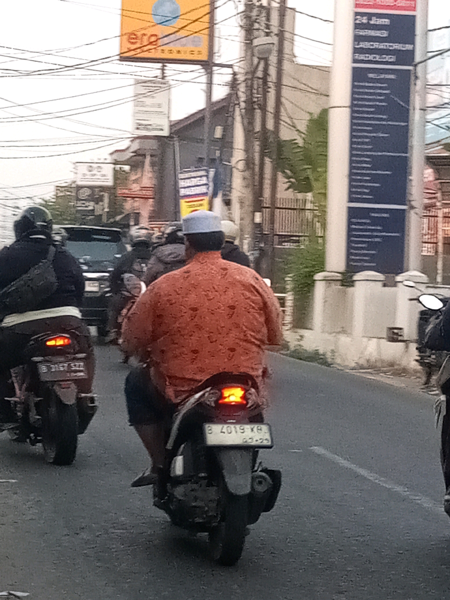 In this scene taken in Bekasi, Indonesia, multiple motorbikes navigate a street. The primary focus is on a man in the foreground riding a motorbike, wearing an orange, patterned shirt and dark shorts. His motorbike's rear license plate reads "B 4019 XB" and "03-29". To his left, another motorbike is visible with a license plate reading "B 3167 SZZ". A black SUV is partially visible in the background, behind other motorbikes. Overhead, numerous electrical wires crisscross the sky. Two billboards are present: one orange and yellow, with the text "ere" and a blue circle, and a tall blue and white sign with text including "24 Jam" and "LABORATORIUM". The environment appears to be a moderately busy urban street.