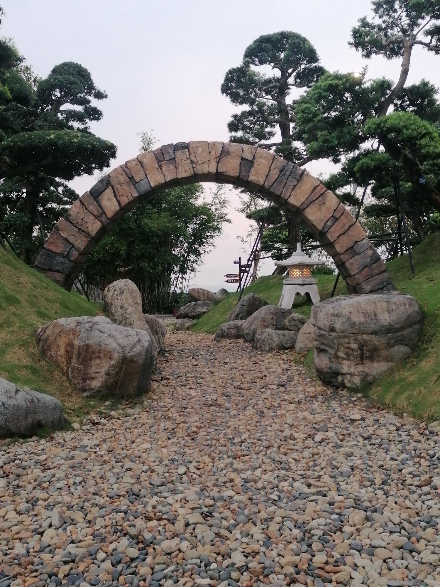 The image depicts a scenic pathway in a park located in Hong Gai Ward, Vietnam. A stone archway frames the entrance to the path, which is covered in small, rounded stones. Large boulders flank the path on either side, leading into a landscaped area with grass and trees. A traditional Japanese-style stone lantern is situated near the archway, providing light. In the background, bamboo stalks and additional signage can be observed. The sky is overcast.