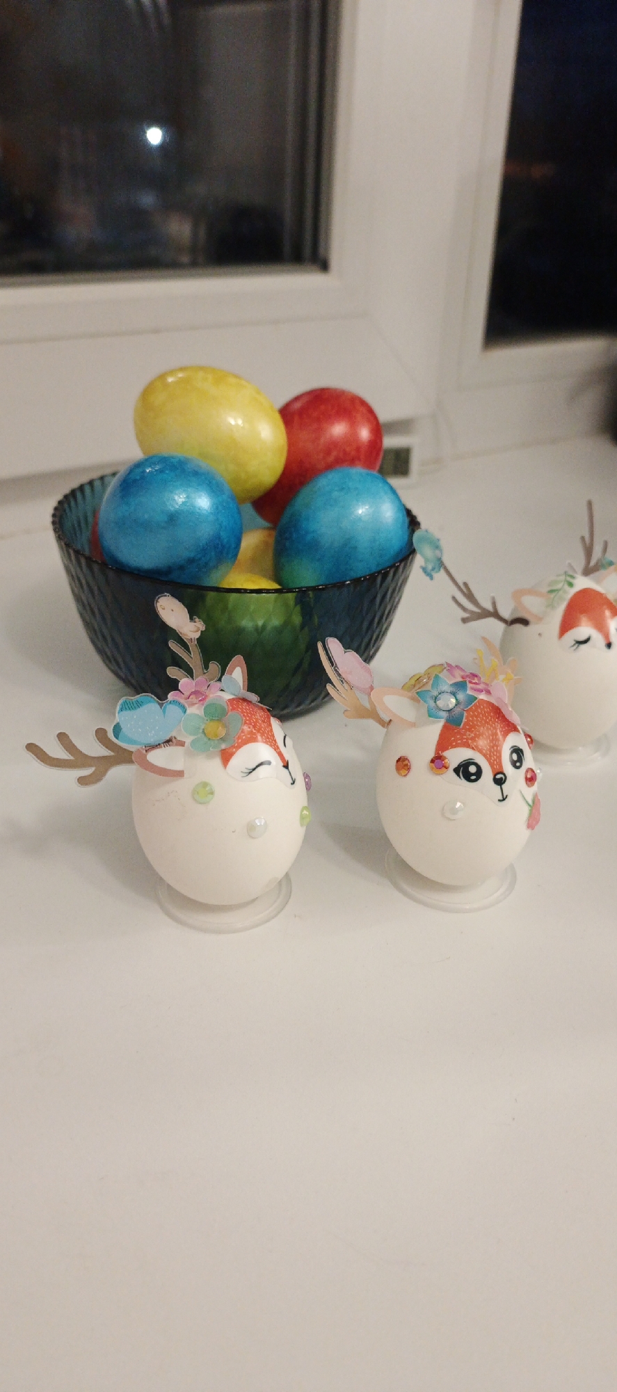 The scene is set on a white surface, likely a table or countertop, near a window. The window is partly visible and appears to be closed with multiple panes. There is a bowl containing several colorful Easter eggs in the background. The eggs are dyed in various colors, including yellow, red, and blue. In the foreground, there are three Easter eggs decorated as deer. Each egg is adorned with a deer face and antlers, along with floral decorations. The decorated eggs are standing on small, clear bases. The exact location in the city of Chaikovsky, Russia, cannot be determined from the image. There are no people visible in the image, only the objects mentioned above.