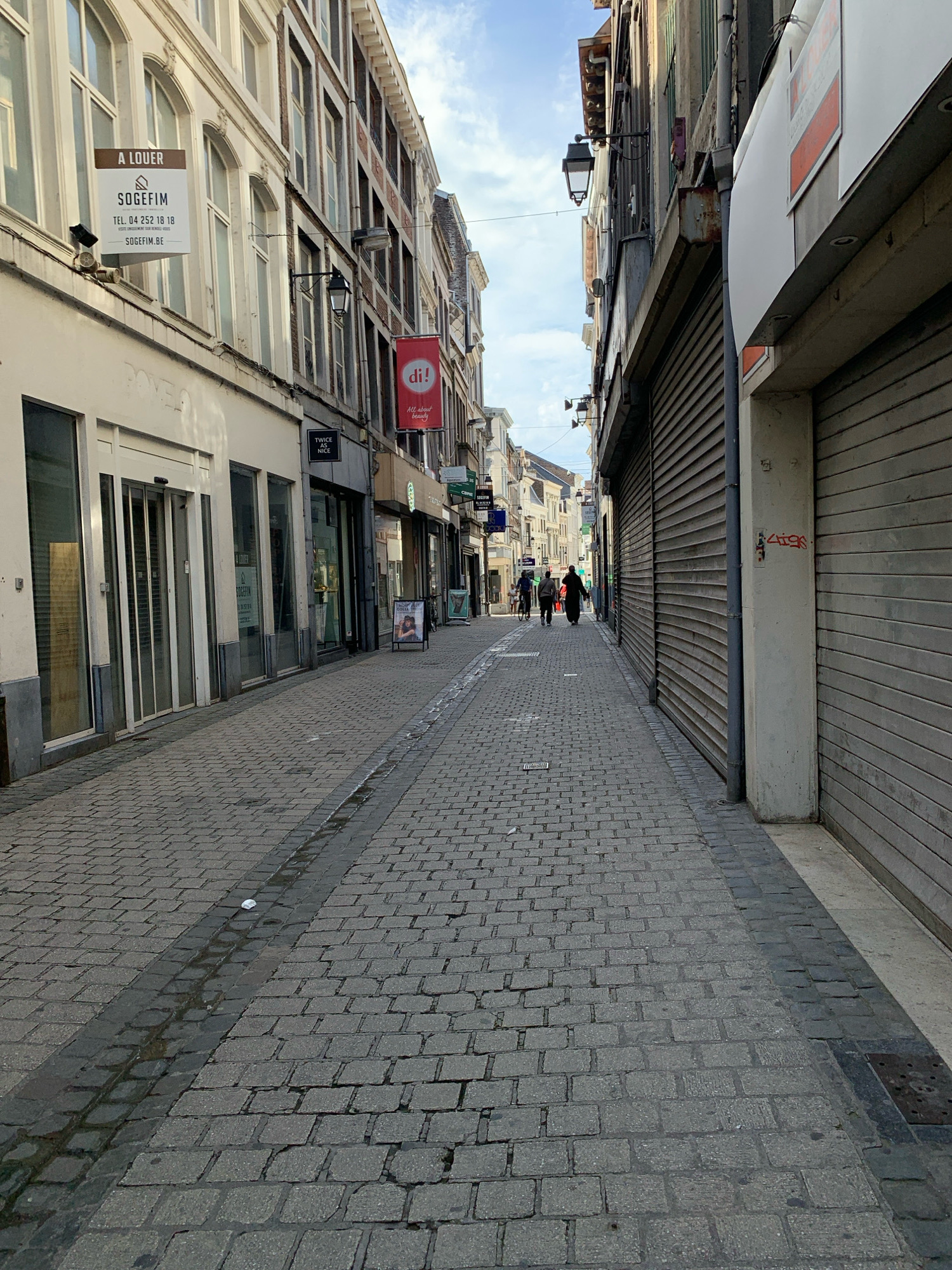 The image depicts a narrow cobblestone street in Liège, Belgium, lined with multi-story buildings. The street is paved with grey cobblestones, with a darker, possibly metallic, strip running down the center, featuring a visible drain cover at the far end. The buildings on either side of the street are a mix of architectural styles.
On the left, the buildings have lighter colored facades with arched windows on the upper floors and storefronts at street level. One storefront has large glass doors and windows. A prominent white sign with the text "À LOUER" (For Rent) and "SOGEFIM" is visible on the building facade. Another building on the left has a red banner with "di!" and "All about beauty" printed on it. Further down the street, several shops appear to be closed, with metallic shutters pulled down.
On the right, the buildings have darker facades and are primarily characterized by large, grey, horizontal roller shutters, suggesting businesses are closed or not yet open. Graffiti, consisting of red spray paint, is visible on a concrete pillar in front of the shutters.
A few people are present on the street. Two individuals are walking away from the camera towards the background, one of them appears to be riding a bicycle. Another two individuals are walking towards the camera in the distance. The sky above is partly cloudy. The overall impression is of a commercial street, possibly in a European city, with many establishments currently closed. The specific location is Rue de la Cathédrale in Liège, Belgium.