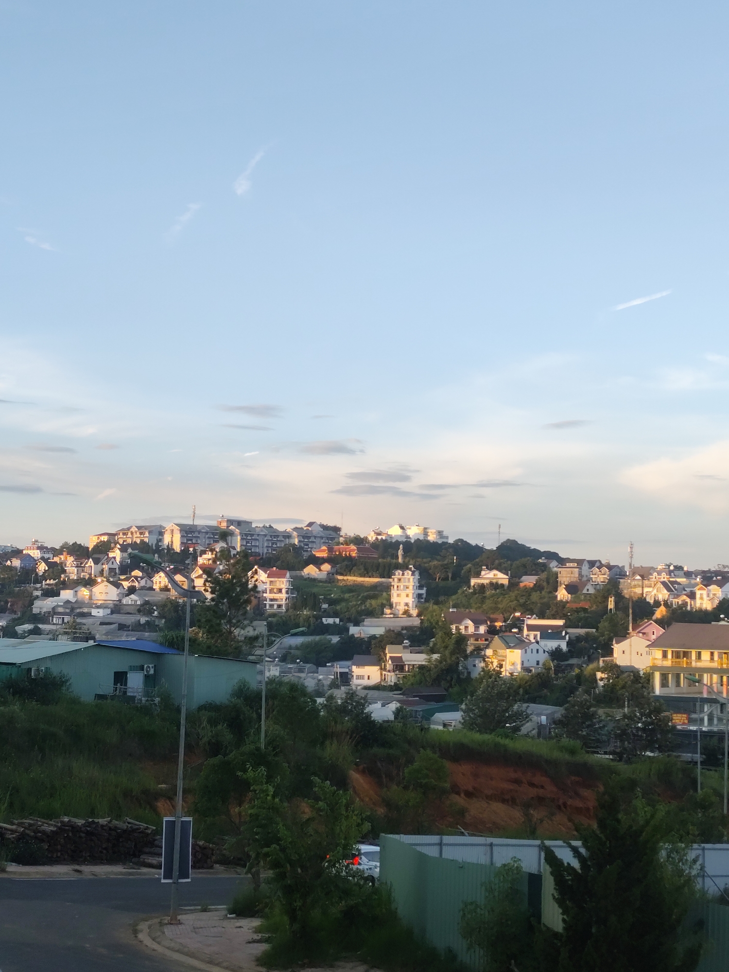 A panoramic view of the city of Da Lat, Vietnam, showcases a dense residential area built on a hillside. The architecture is varied, with a mix of multi-story buildings and smaller houses, many featuring colorful roofs and balconies. Vegetation is abundant, with trees and shrubs integrated into the urban landscape, and a prominent green hill slopes down in the foreground. The sky is a soft blue with wispy clouds, suggesting either early morning or late afternoon light, as evidenced by the warm glow on some of the buildings. A road curves through the bottom of the frame, bordered by a green fence. There are no visible people in the image.