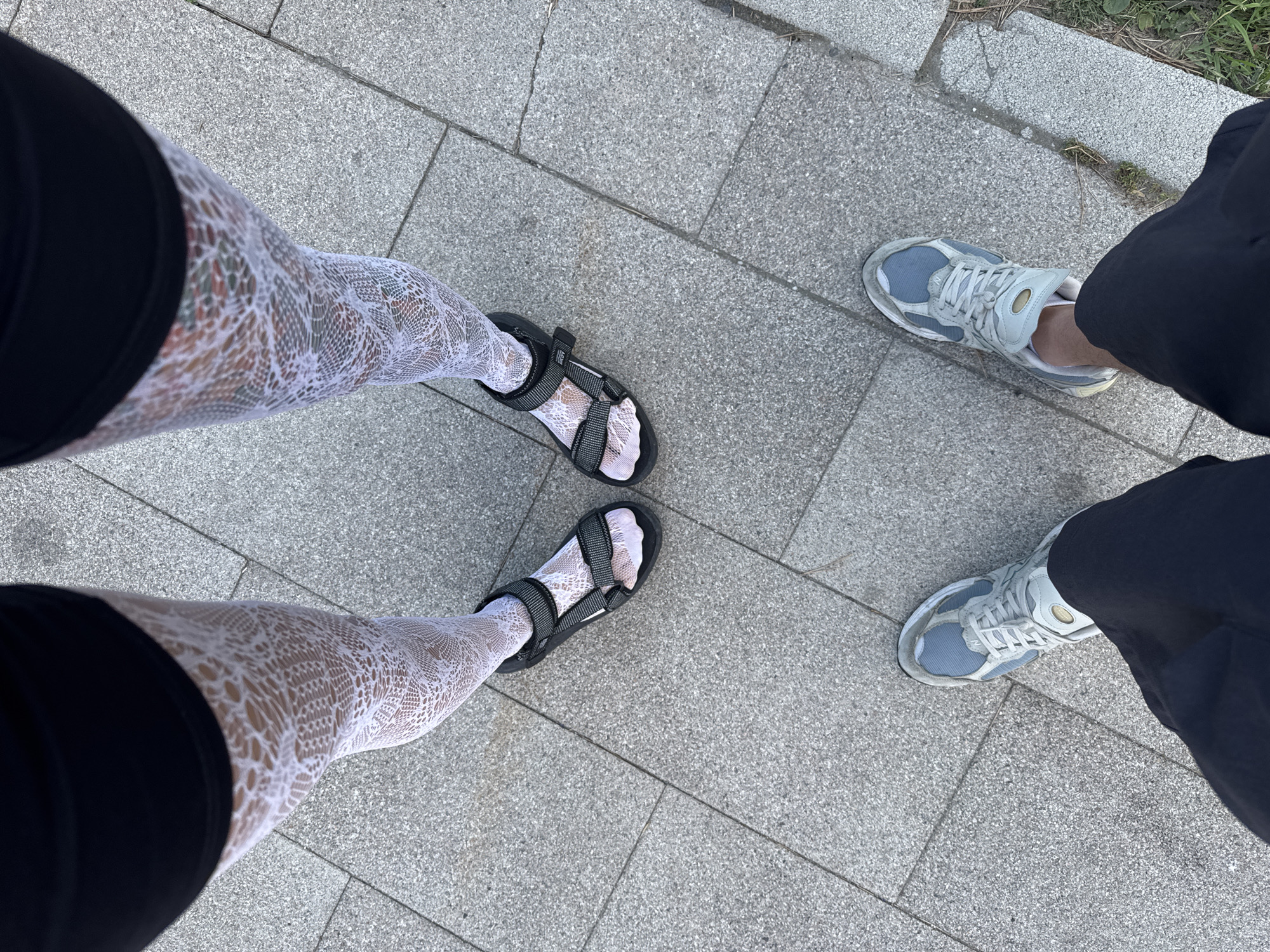 The image shows two pairs of legs and feet standing on a paved surface, likely a sidewalk or plaza. The left legs are wearing white lace stockings and black sandals, while the right legs are wearing dark pants and light blue and grey sneakers. The pavement is made of large, grey, rectangular tiles. In the upper right corner, there is a hint of green grass and dry vegetation, suggesting an outdoor setting. The lighting indicates it is daytime, likely a clear day given the shadows. The scene appears to be a casual outdoor moment, possibly during a walk. There is no visible text in the image. The context indicates the location is Moscow, Russia.