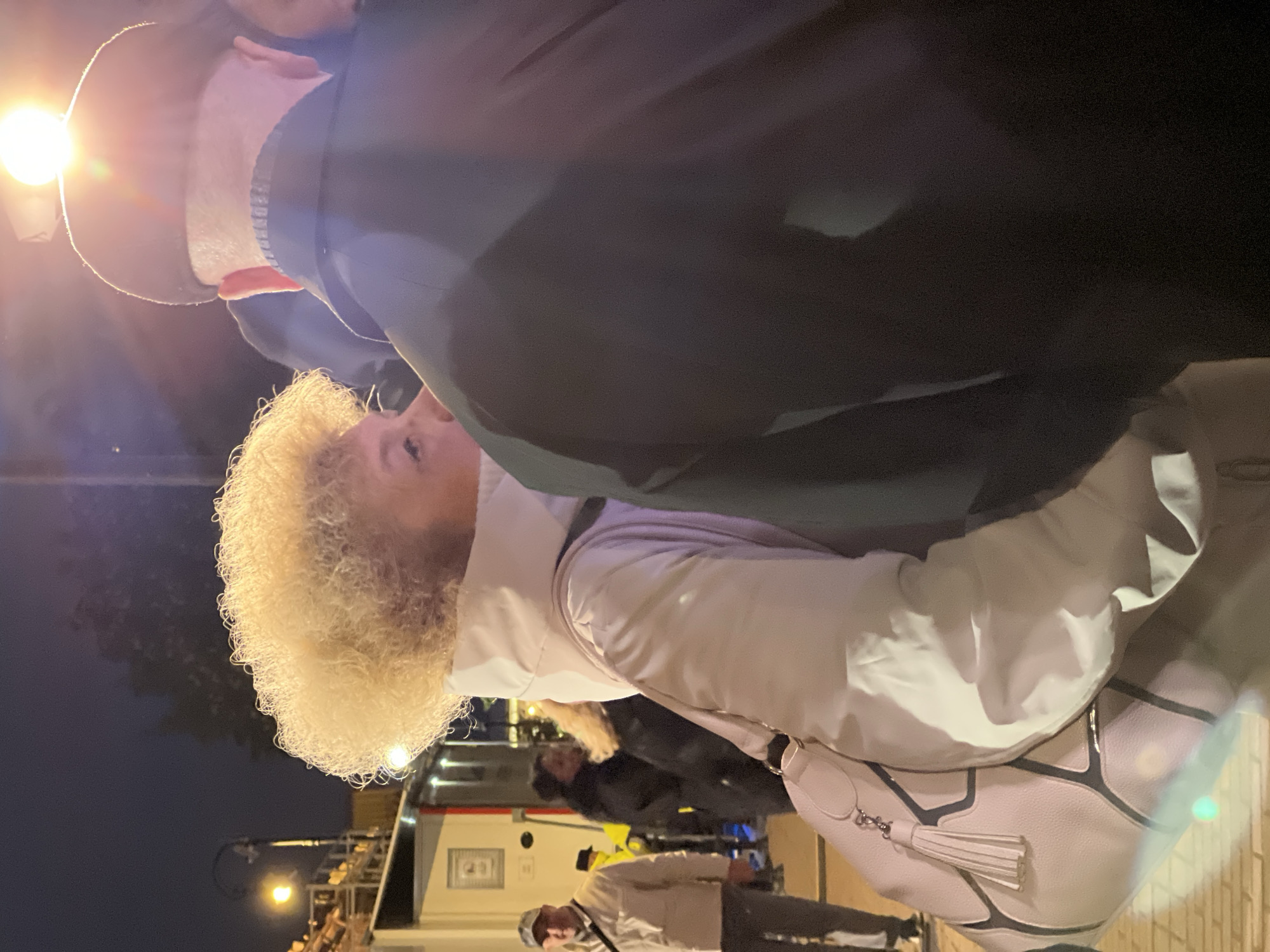 The image shows a group of people at night in Saint Petersburg, Russia. A woman with blonde curly hair is in the foreground wearing a light-colored jacket and a white bag. Behind her is a man in a black coat. In the background, there is a bus and other people, including one man wearing a light-colored jacket and a baseball cap. Streetlights are visible, and the sky is dark.