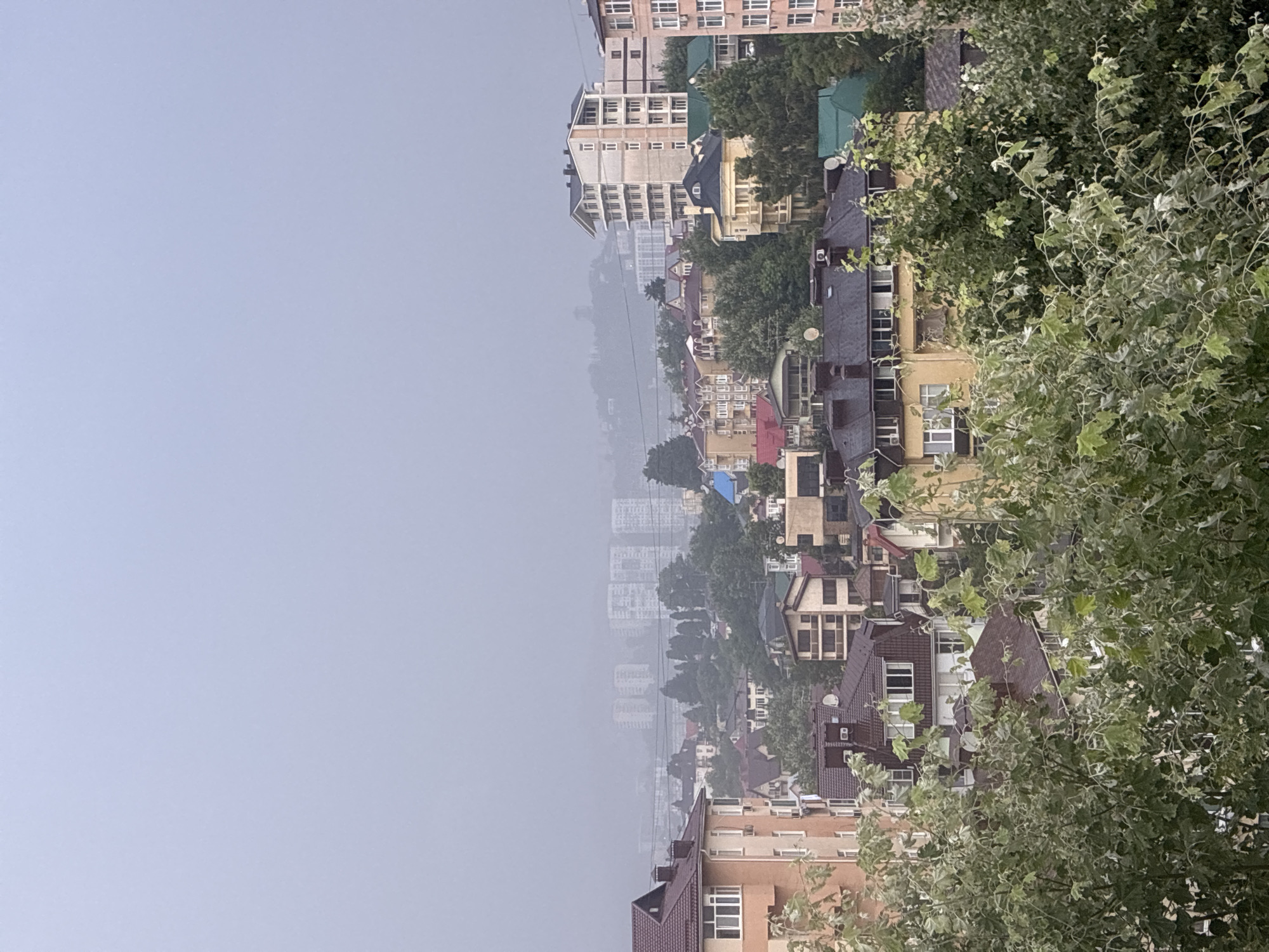 The image captures a hazy, overcast view of a densely populated urban area with a mountainous backdrop, likely in Sochi, Russia. In the foreground, lush green foliage obscures the lower portion of the scene. Above the foliage, a cluster of buildings of varying architectural styles and heights is visible. These include multi-story residential buildings with tiled roofs in earthy tones, as well as taller, more modern apartment complexes in shades of white and beige. Some buildings feature balconies, window units, and satellite dishes.
Further into the background, more structures are discernible through the atmospheric haze, including what appear to be larger apartment blocks and possibly some industrial buildings. The distant landscape is shrouded in a dense mist, softening the outlines of the terrain and obscuring any fine details. The overall impression is one of a coastal city nestled among hills, experiencing a day with low visibility due to fog or heavy cloud cover. No people or specific activities are evident in the image.