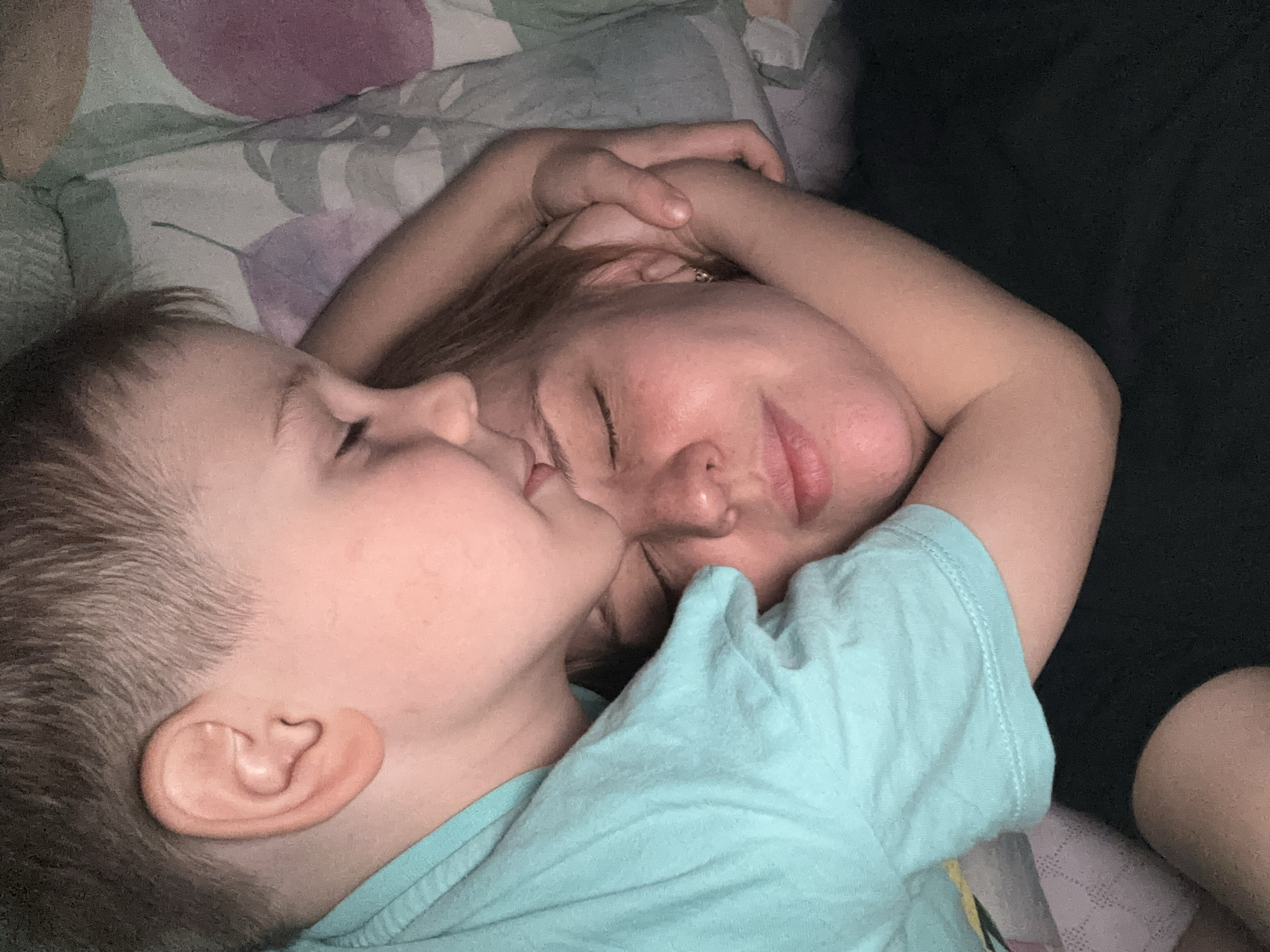 A young child is nestled beside a woman, both lying down. The child's face is close to the woman's, as if kissing her cheek or nuzzling her face. Both have their eyes closed. The location and city of Rusanovo, Russia are not confirmed, since they are absent from the image.