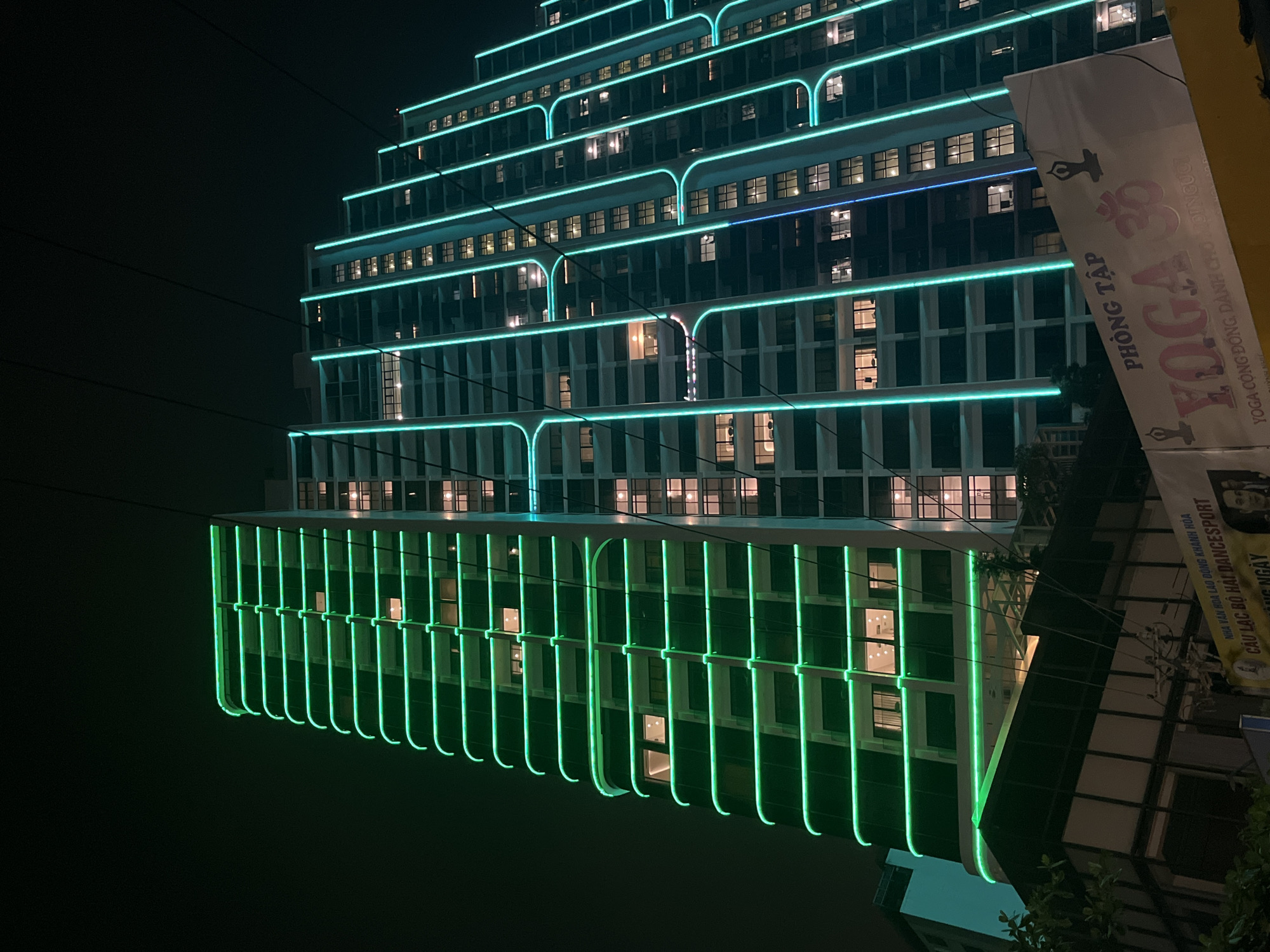 The image captures a nighttime scene in Bac Nha Trang Ward, Vietnam. The focal point is a modern high-rise building adorned with vibrant green neon lights accentuating its unique stepped architecture. The building's design features horizontal neon bands that extend vertically between the windows, creating a grid-like pattern. The building is illuminated with internal lights visible through the windows. Partially obstructing the view is a sign, printed in Vietnamese, advertising a Yoga studio, positioned in the foreground, with text: "PHÒNG TẬP YOGA...YOGA CỘNG ĐỒNG DÀNH CHO MỌI NGƯỜI". The surrounding environment is dark, implying a night setting. A few power lines are visible in the upper part of the image. Overall, the scene is illuminated by the building's neon lights and street lighting, casting a vivid and contemporary ambiance.