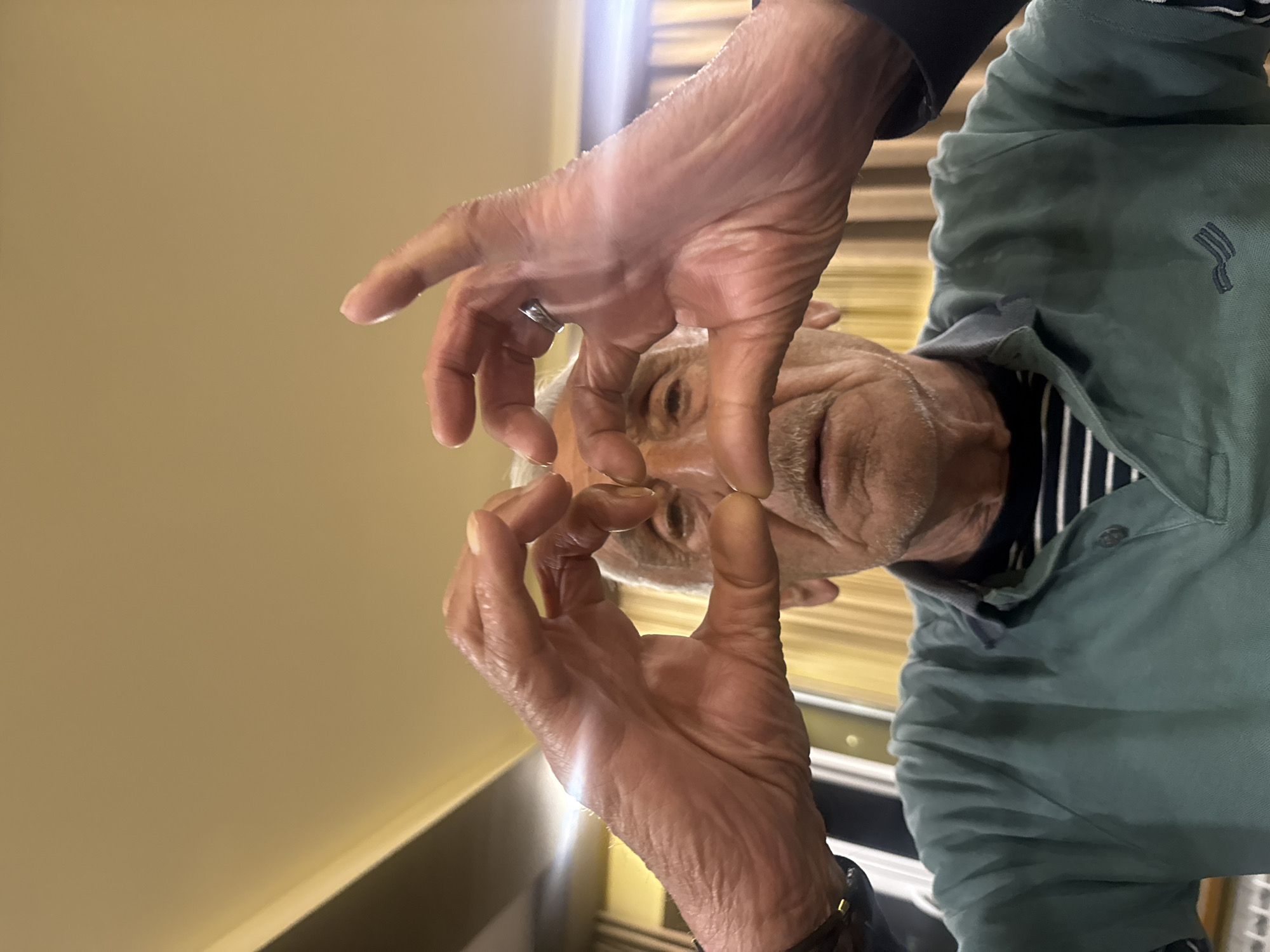 A close-up shot shows an older man with white hair and wrinkled skin, making a small circle with his thumb and forefinger, framing his face. He wears a green button-down shirt with a navy blue and white striped undershirt. A ring is visible on his left ring finger and a watch on his left wrist. The background consists of a light-yellow wall on the left and a wooden-slatted blind or wall panel on the right. The environment does not explicitly indicate the location to be Yerevan, Armenia.