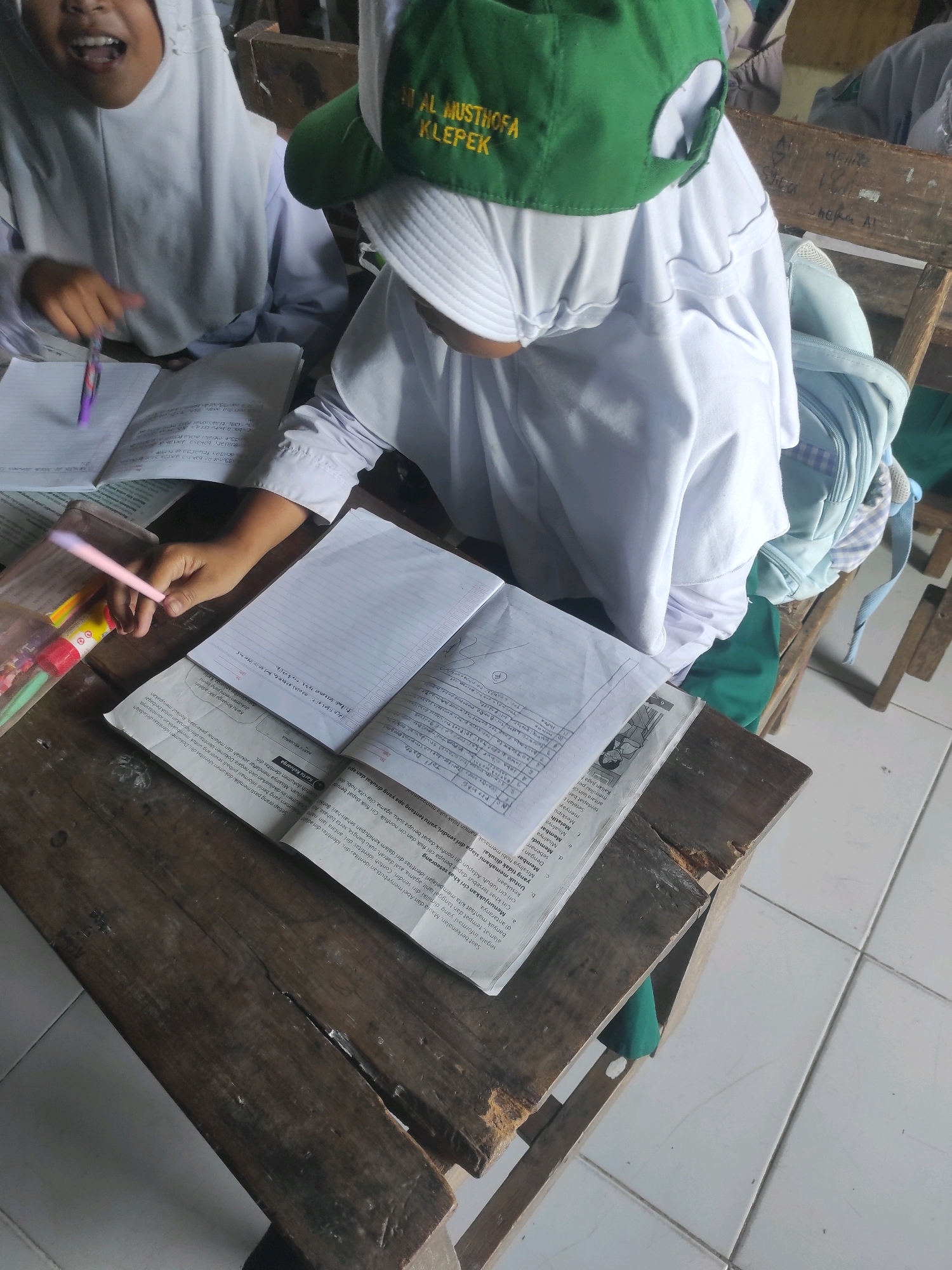 Two young female students, likely of elementary or middle school age, are seated at a wooden desk in a classroom setting. Both are wearing white hijabs and uniforms. The student on the left has her mouth open in a surprised or excited expression and is holding a purple pen. The student on the right is wearing a green baseball cap with yellow lettering that reads "AL MUSTHOFA KLEPEK". Her face is partially obscured by the cap and hijab, but she appears to be looking down at her work.
In front of the students are several open books and notebooks. One notebook is open to a page with handwritten notes, and another is a printed textbook with dense text and a small illustration. A pencil case filled with colored pens and pencils sits to the left of the notebooks. A light blue backpack with a checkered pattern is visible behind the student on the right.
The desk is made of rough, weathered wood, with visible cracks and imperfections. The classroom environment appears basic, with other wooden desks and chairs visible in the background. The floor is tiled in a white checkerboard pattern. The overall scene suggests a typical school day in a classroom in Klepek, Indonesia, with students engaged in their studies.