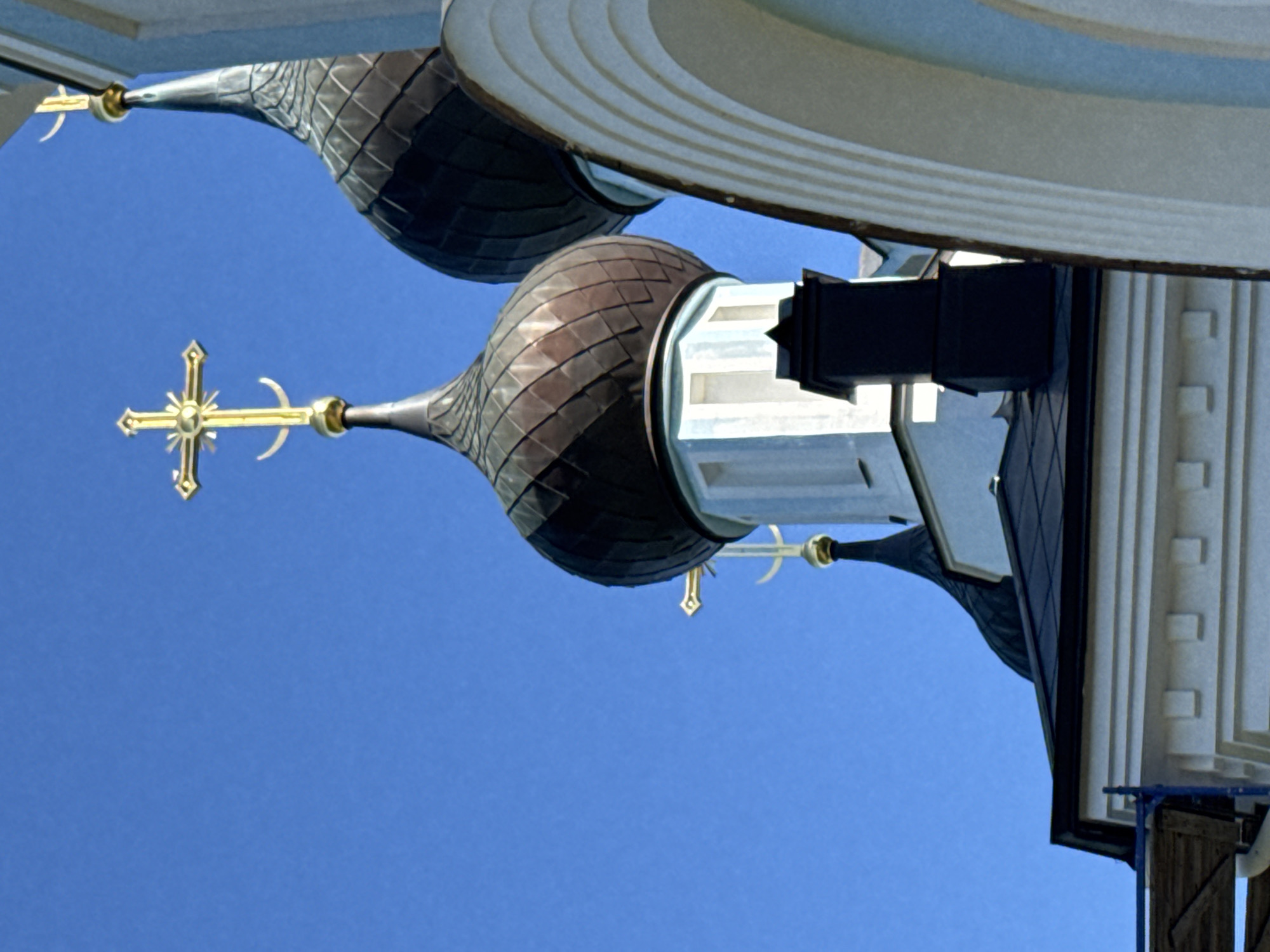 The image shows the domes and crosses of a Russian Orthodox church, seen against a clear blue sky. The architecture features multiple onion domes, one of which is a prominent dark, metallic color, while another is lighter and features arched architectural elements. Golden crosses, characteristic of Orthodox churches, are mounted atop the domes. One cross is particularly detailed with a circular element and a crescent moon shape at its base. The church building itself appears to be white with light blue accents. A black chimney or vent is visible on one of the structures. The scene is a detail-oriented shot, focusing on the religious architecture. The location is identified as St. Ozerki, Russia. There are no people or animals depicted in the image.
