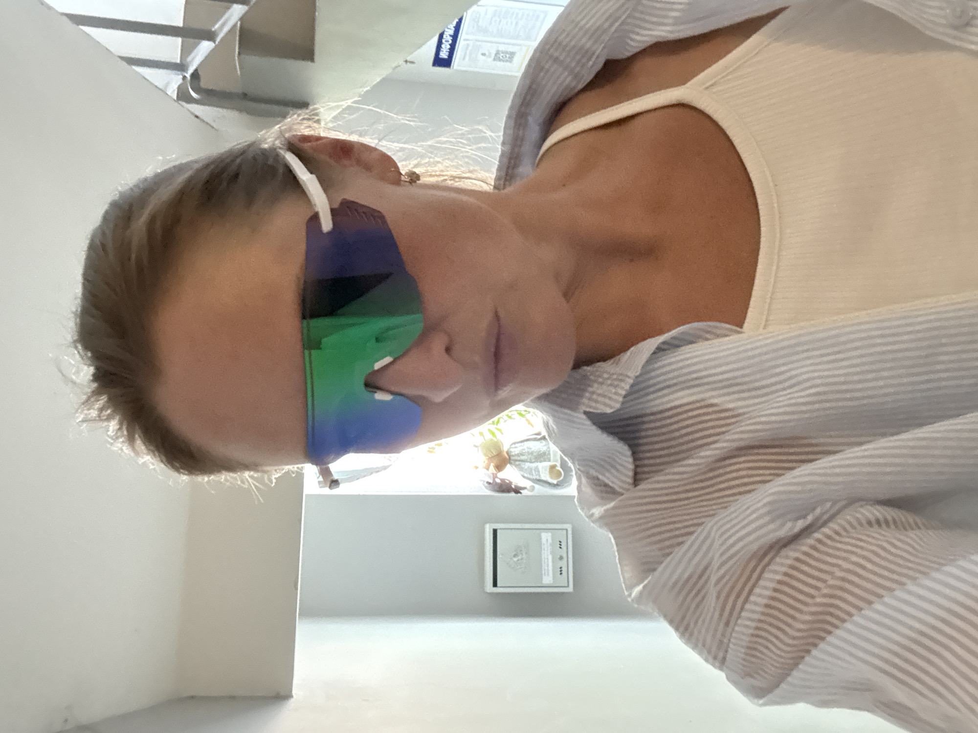 A woman with fair skin and blonde hair is wearing blue and green sunglasses, a white tank top, and a light blue and white striped shirt. She is indoors, possibly in the city of Chaikovsky, Russia, though this cannot be confirmed. A framed picture is on the wall of the room. There is a window in the background. The image is taken from a low angle looking upward.