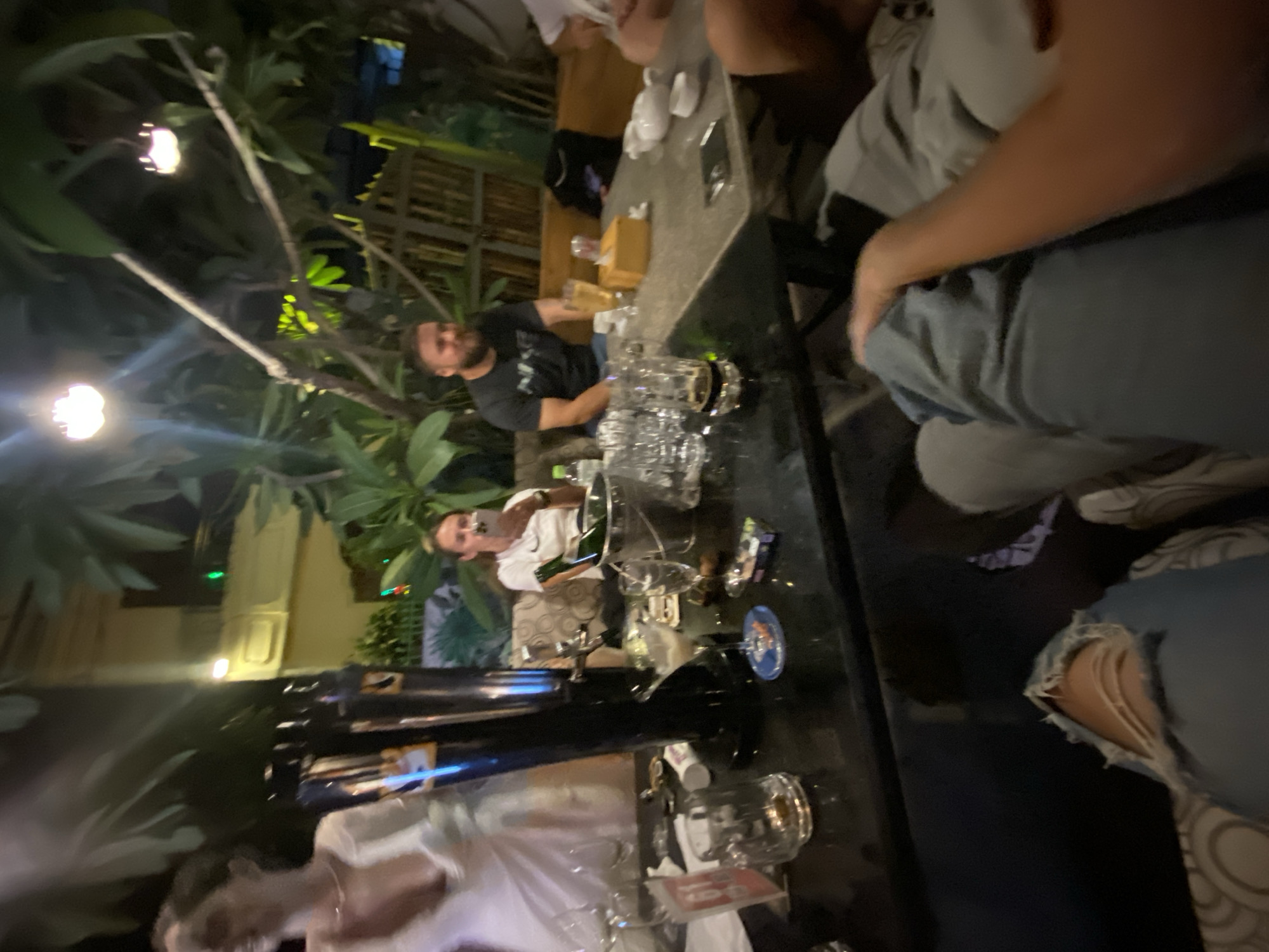 This is a medium shot, indoor, eye-level, candid photograph taken at night in Phu Thuy Ward, Vietnam. A group of people are gathered around a table, appearing to be at an outdoor bar or restaurant. In the foreground, a person's torn blue jeans are visible, indicating they are sitting down. To the right, another person's arm rests on a lap, and to the left, a woman in a white dress is partially visible.
In the midground, a man with a beard wearing a black t-shirt is seated at the table, looking to his left and smiling. To his left, a woman in a white t-shirt is holding a mobile phone to her ear. Both individuals appear to be engaged in conversation or observing something. The table is cluttered with various objects, including glasses, bottles, an ice bucket with a bottle of champagne, and a tissue box. A prominent black beer tower with a tap is positioned on a side table.
The background reveals a building with yellow walls and a dimly lit interior, partially obscured by lush green foliage and trees. String lights are visible, casting a warm glow and contributing to the nighttime ambiance. The overall scene suggests a casual and convivial gathering.