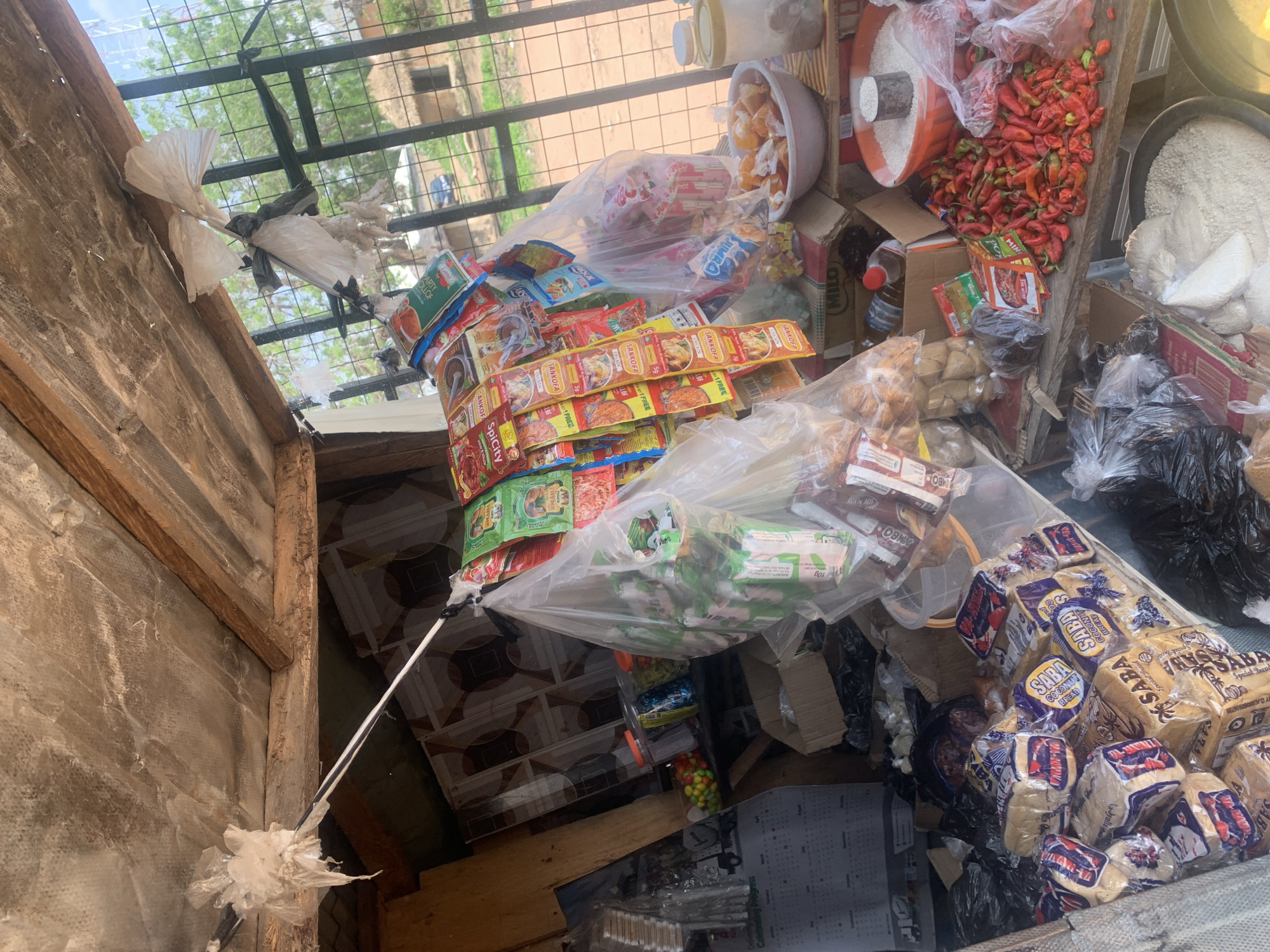 This is a high-angle, eye-level shot of a cluttered outdoor market stall, possibly in Jalingo, Nigeria. The scene is densely packed with various goods, suggesting a busy trading environment.
Prominent objects include stacks of packaged bread, identified by the "SABA" branding, as well as numerous small packets of spices and seasonings, many labeled "SANKOFA" with varying flavors like "CHICKEN" and "SPICY CITY." There are also bags of rice, a large quantity of red chili peppers, and what appear to be snacks or candies in jars. A few bottles of oil and containers of Milo are also visible.
The stall itself is constructed from rough, weathered wood and covered with a metal grate or fencing overhead, through which trees and a glimpse of the sky can be seen. The ground appears to be tiled in a geometric pattern. The overall impression is one of a vibrant, somewhat disorganized marketplace.
The lighting suggests it's daytime, likely morning or afternoon. There are no people clearly visible in the frame, but the abundance of goods implies a commercial activity. Several white plastic bags are tied to a wire or string, adding to the chaotic yet purposeful arrangement of the stall. The text on the packaging is mostly in English, with some possibly in local languages, indicating commercial products for sale.