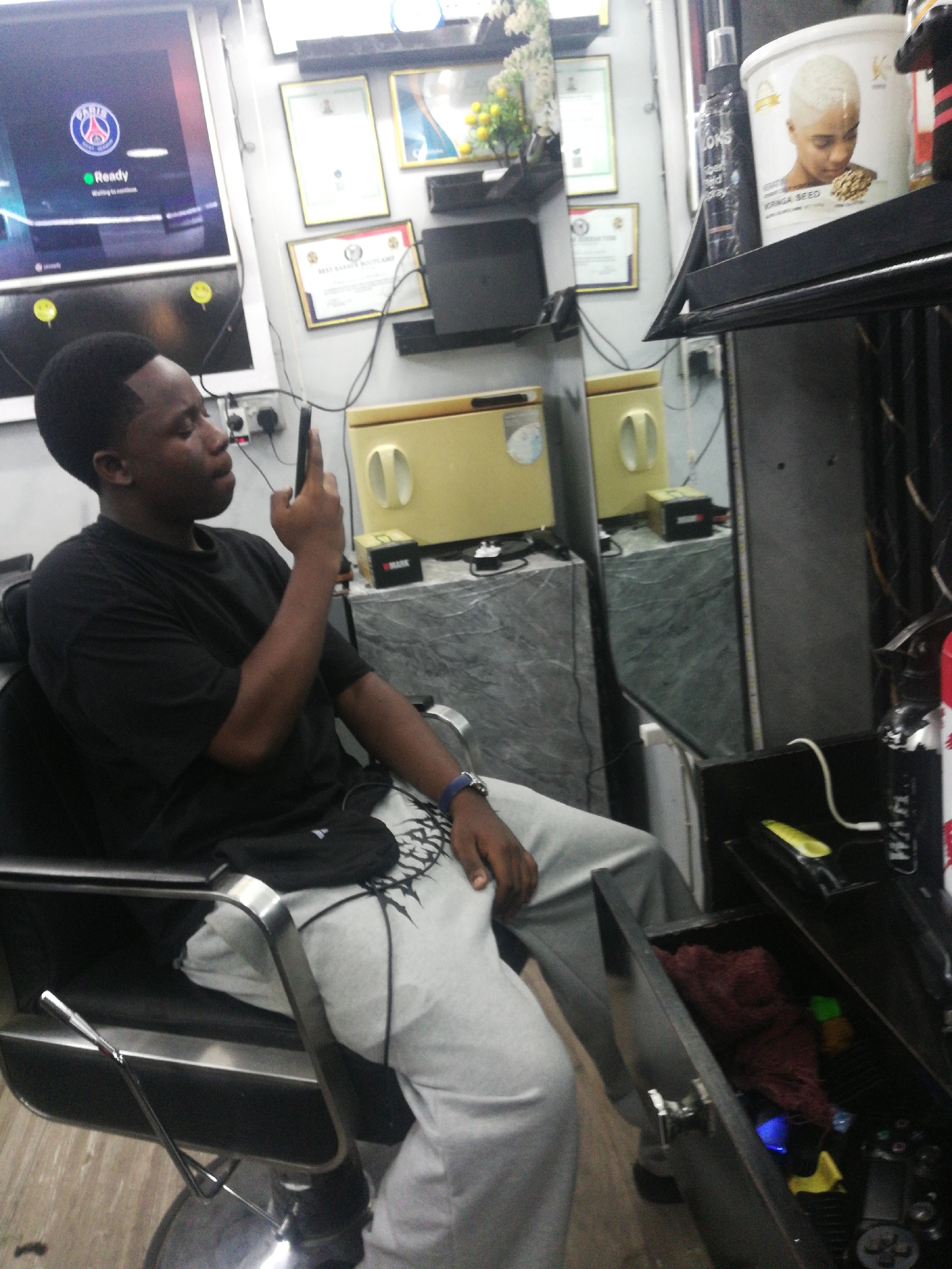 The image captures a young man sitting in a barber chair in what appears to be a barbershop setting, potentially in Lagos, Nigeria. The man is wearing a black t-shirt and grey sweatpants, and he is looking at his phone. The shop features a flat-screen TV showing a video game, framed certificates on the wall, barber's tools, and hair product containers on shelves. The environment appears to be a busy barbershop interior.
