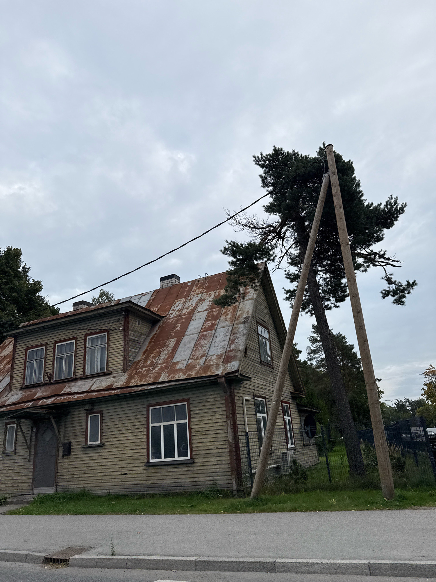 The image shows a weathered, two-story wooden house with a rusty corrugated metal roof. The house is located on a street in Tallinn, Estonia. Two large wooden poles are propped against a tall pine tree next to the house, suggesting some form of temporary support or construction. The house has multiple windows, some with white frames and others with red trim. A dark gray door is visible on the left side of the house. The surroundings include a patch of grass in front of the house and a dark fence in the background, behind which more trees and some structures are visible. The sky is overcast and gray.