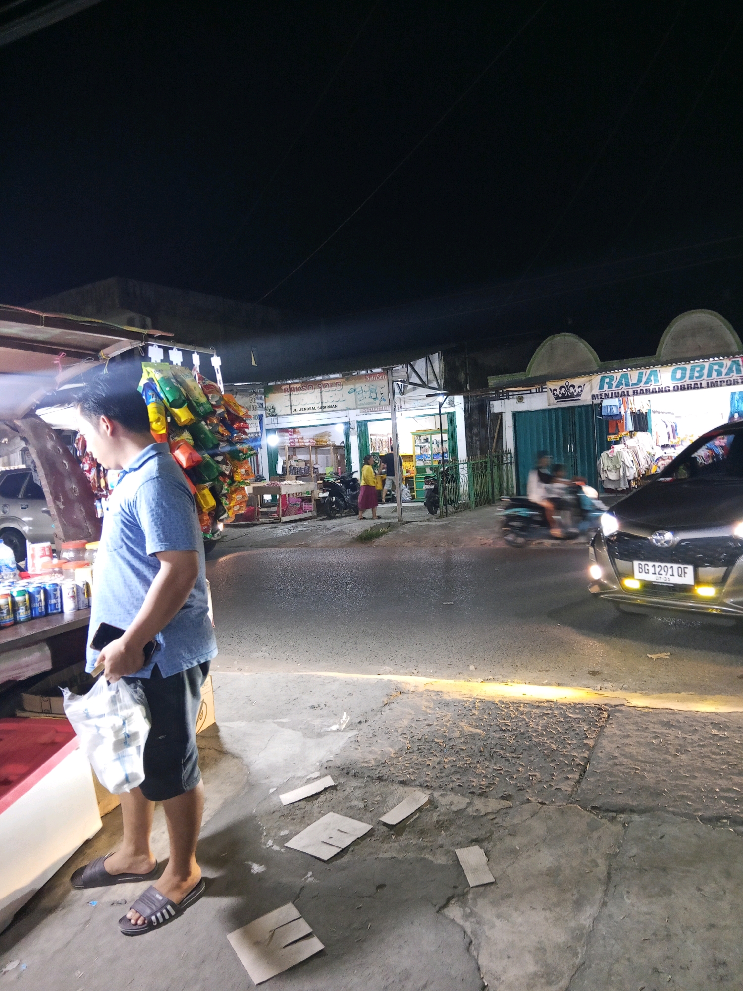 The image depicts a street scene at night in Lubuklinggau, Indonesia, likely in front of a row of shops. A man in a blue polo shirt and dark shorts stands on the sidewalk, holding a plastic bag and a mobile phone. He is facing away from the camera, looking towards a street stall laden with packaged snacks. To the right, a dark car with its headlights on is parked on the road, displaying the license plate BG 1291 QF. In the background, a building with a sign that reads "RAJA OBRA" is visible, suggesting it is a shop or establishment. Other individuals and motorcycles are present in the distance, indicating a lively urban environment. The ground is paved with concrete and shows signs of wear and tear, with scattered debris. The overall lighting suggests artificial illumination from streetlights and shop fronts.