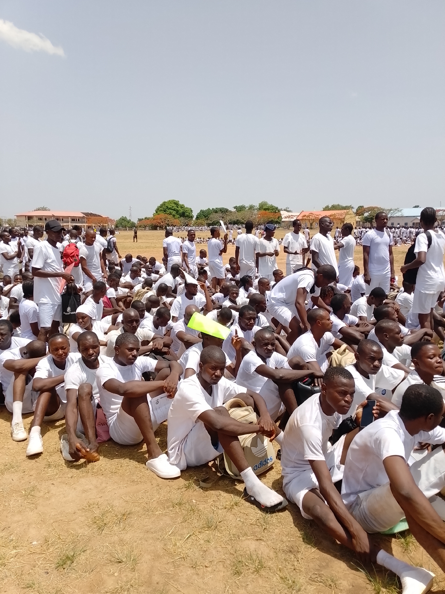 The image displays a large outdoor gathering of numerous young to middle-aged adults, predominantly men, seated closely together on a dry, dusty dirt field with sparse patches of grass. The participants are uniformly dressed in white t-shirts, white shorts, and mostly white footwear, suggesting their involvement in a structured event such as a recruitment drive, orientation, or training camp. They are seated in rows, appearing calm and patient, some looking forward while others engage quietly with those nearby.
In the background, several one-story institutional buildings with light-colored walls and reddish-brown roofs are visible, interspersed with green and reddish-brown trees. The sky above is bright and mostly clear, with a single wispy white cloud, indicating a sunny, mid-day setting. The bright light casts short or indistinct shadows, typical of overhead sun.
Notable objects among the crowd include a black baseball cap worn by a standing individual, various backpacks (one prominently featuring an "Adidas" logo), mobile phones, and a black tote bag with the "AquaFlask" logo. A bright yellow or green folder/document is visible among the seated individuals, and one person in the foreground holds a document displaying the number "10." The overall scene conveys an impression of an organized and disciplined assembly taking place in a warm, outdoor environment.