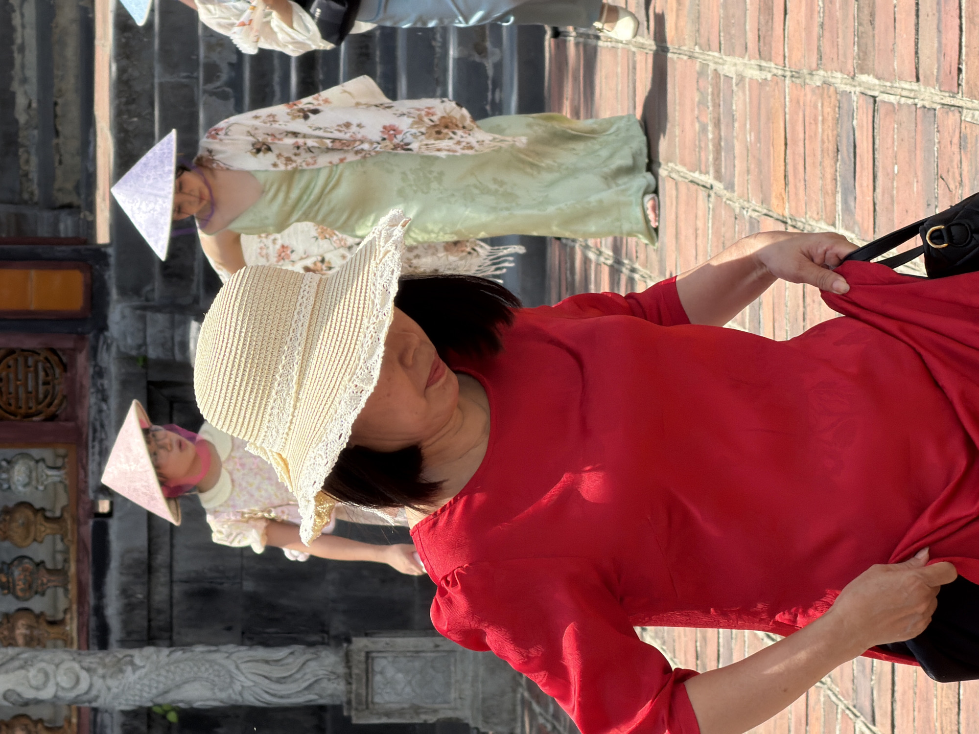 In Phú Xuân Ward, Vietnam, a woman in a red dress and a straw hat walks on a brick pathway. Behind her, two women in conical hats, one wearing a light green dress and a floral shawl, and another in a pink conical hat, stand on stone steps. The background features architectural elements consistent with historical Vietnamese structures, including ornate railings and stone pillars. The lighting suggests it is daytime, with sunlight casting shadows.