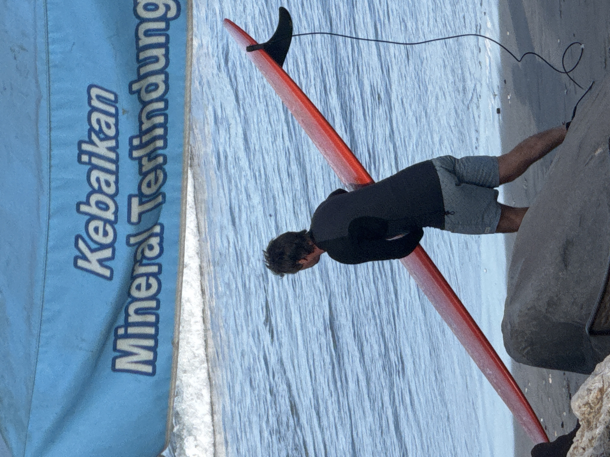 The image shows a person on the beach in Kuta, Indonesia, holding a red surfboard. The person is wearing a black rashguard and grey shorts, and appears to be looking out at the ocean. In the background, a blue banner with white text in Indonesian reads "Kebaikan Mineral Terlindung". The ocean is calm with small waves, and the beach is sandy with some rocks in the foreground. The overall scene suggests a surfing activity or preparation for surfing at a beach known for its surfing spots.
